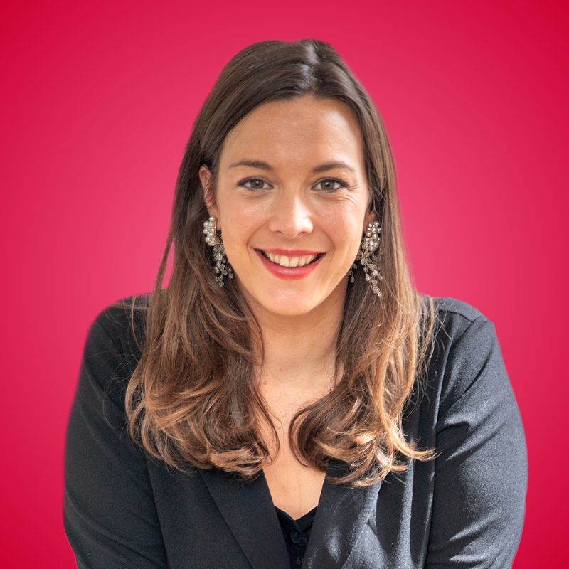 Smiling young woman with long brown hair and pearl earrings poses against a vibrant red background in a dark blazer.