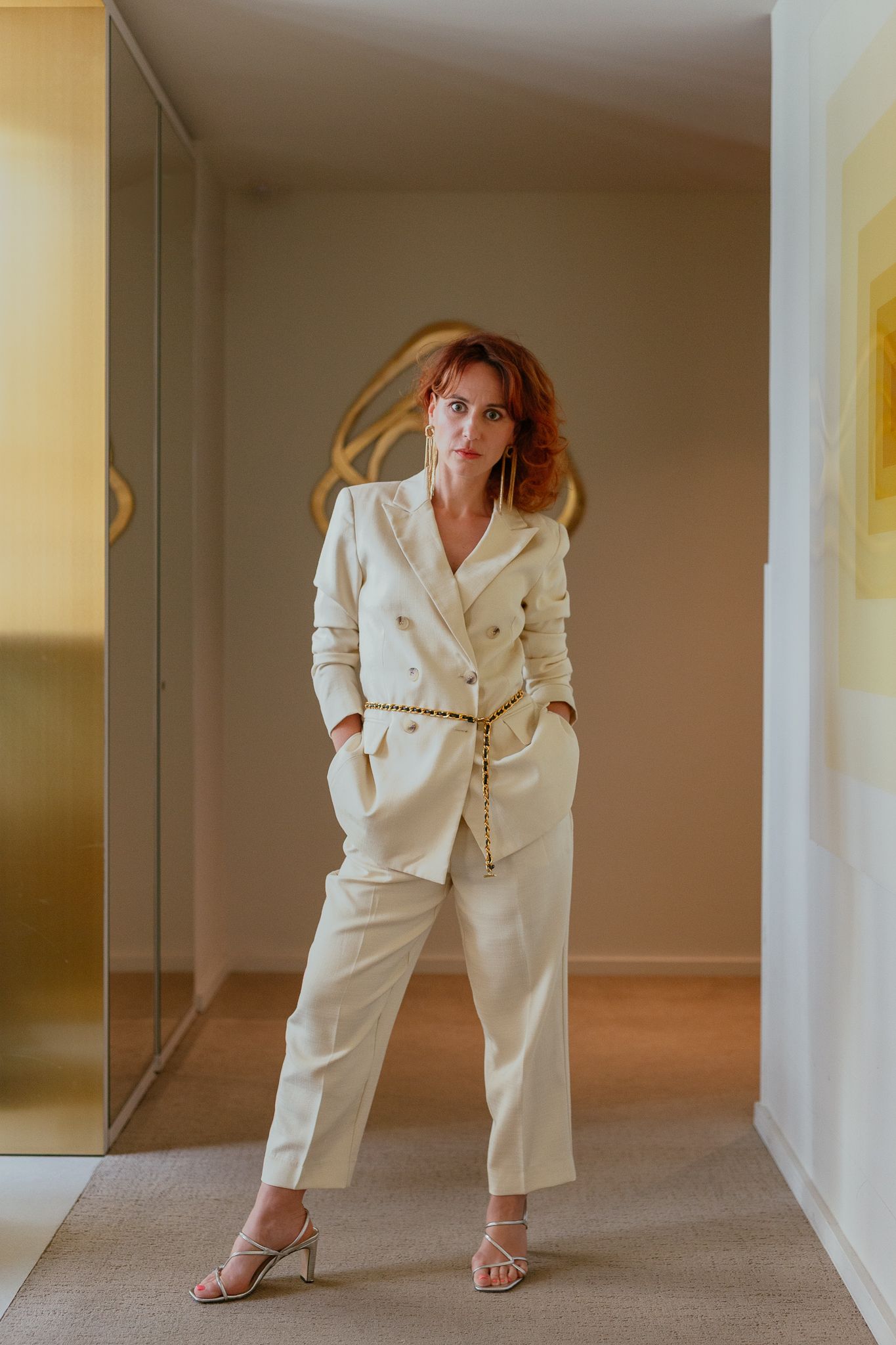 Woman in cream double-breasted suit with gold belt and strappy heels poses in modern interior hallway.