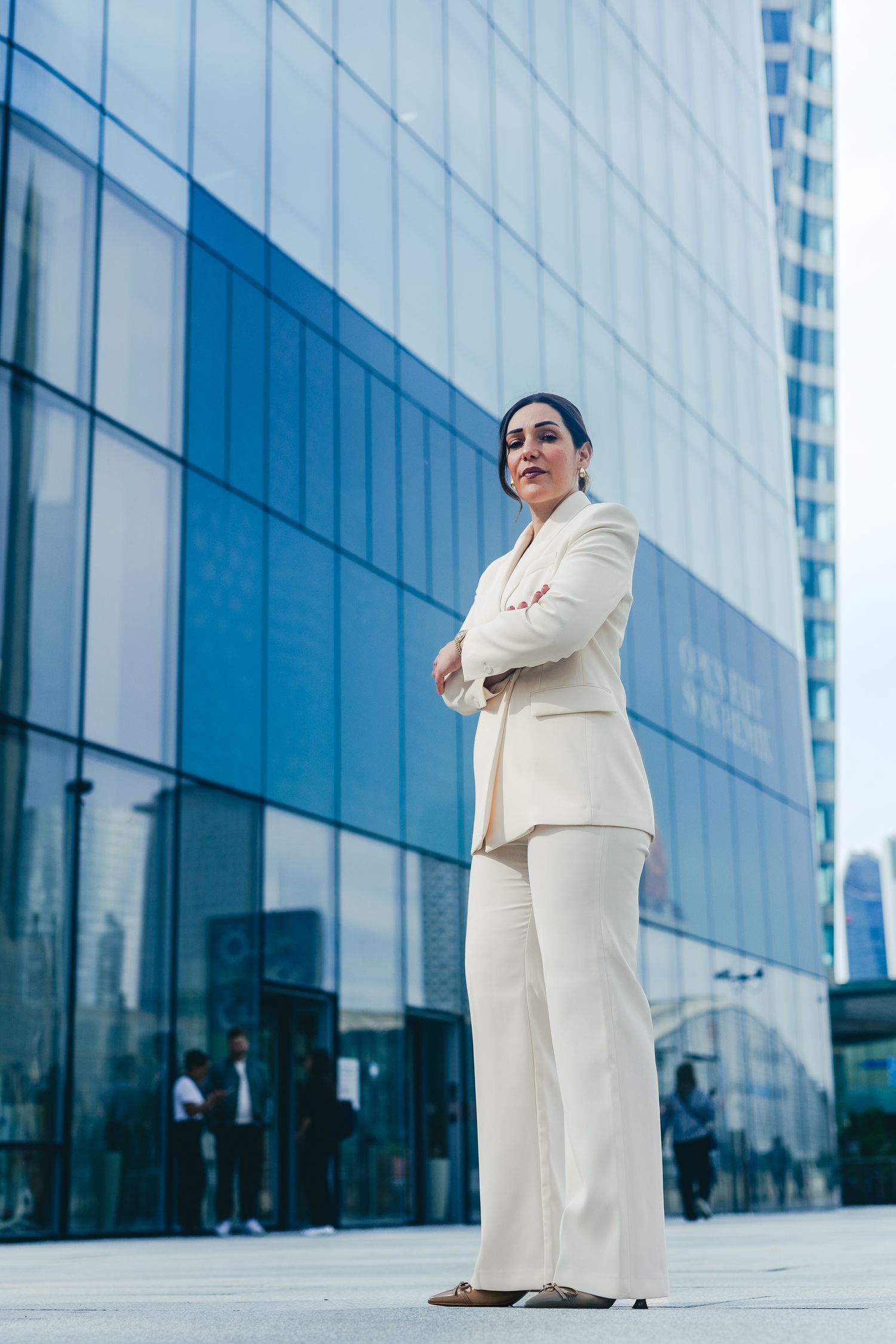 Confident businesswoman in white suit standing arms crossed outside modern glass office building.