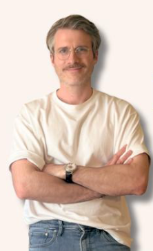 Confident man with mustache and glasses in white t-shirt standing with arms crossed against light background.