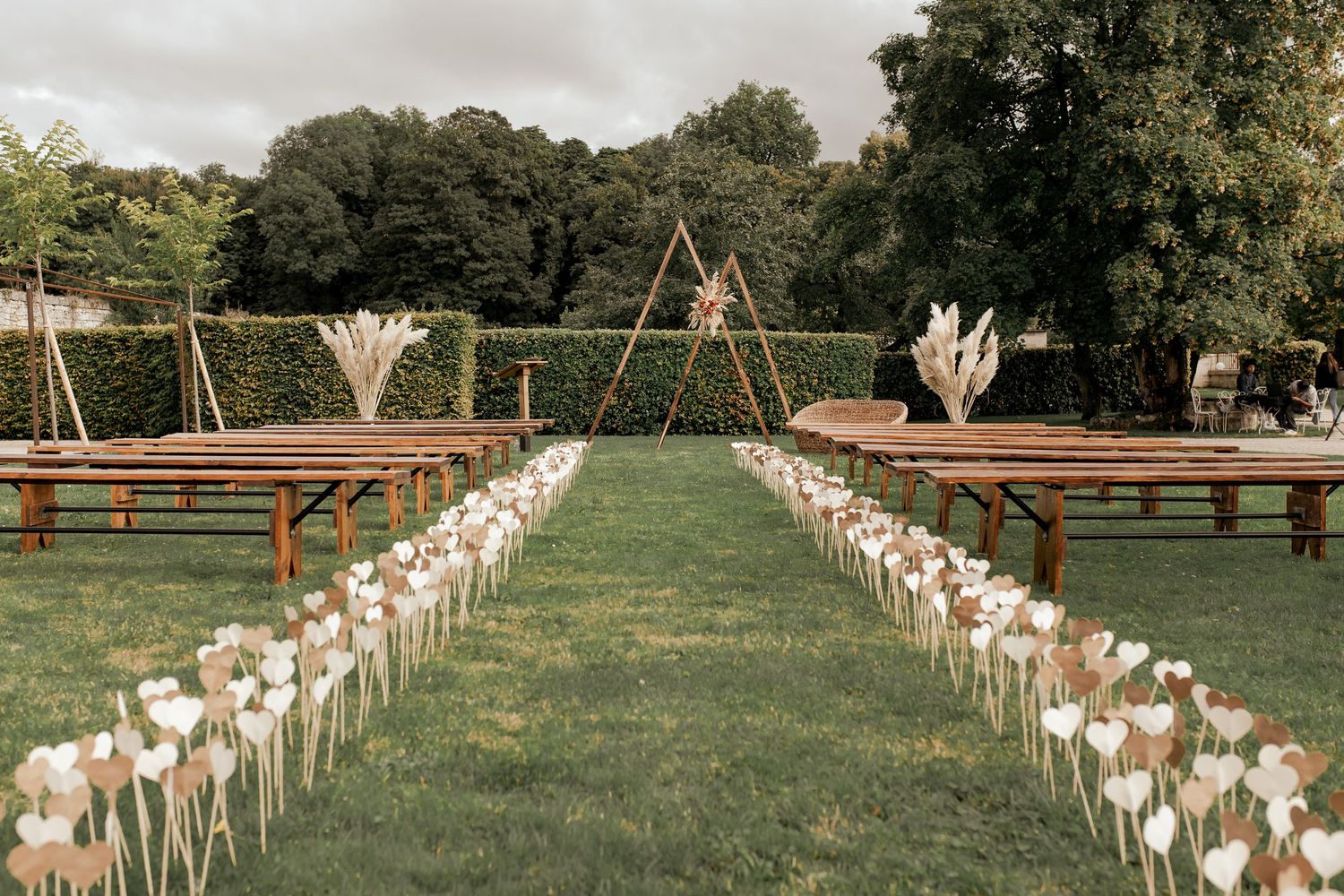 Outdoor wedding ceremony aisle lined with white tulips and pampas grass, wooden benches, triangular arch backdrop on lush green lawn.