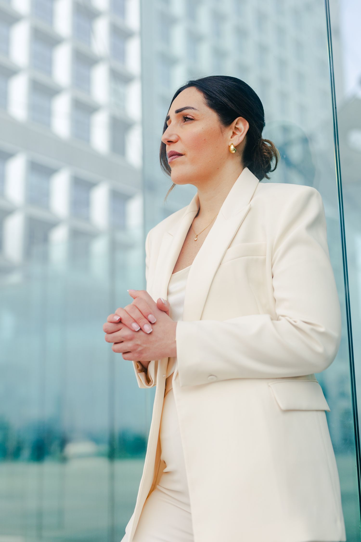 Professional woman in white blazer stands by glass wall with hands clasped in an urban business district.
