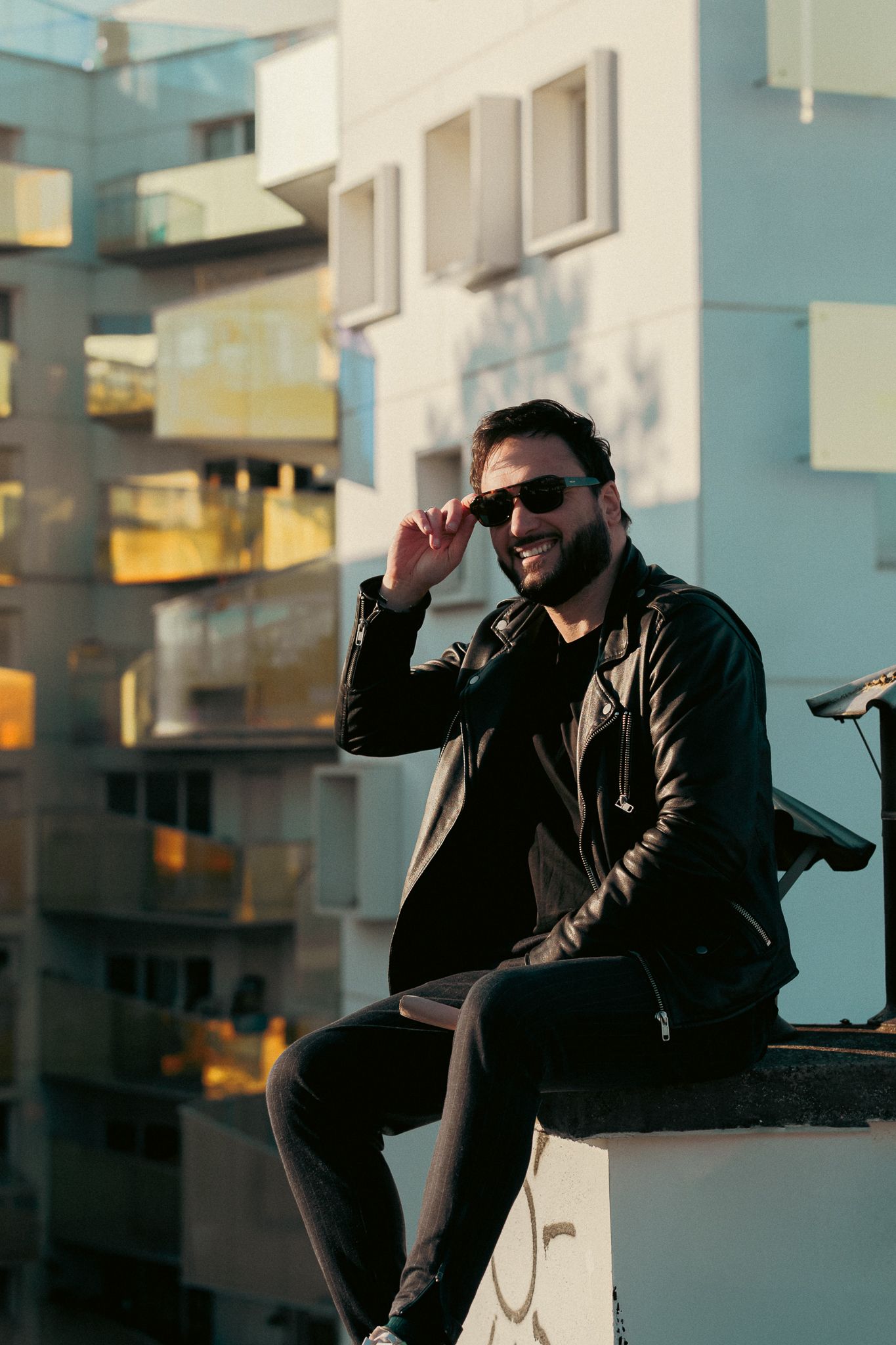 Smiling bearded man in black leather jacket and sunglasses sitting on rooftop with urban apartment building backdrop.