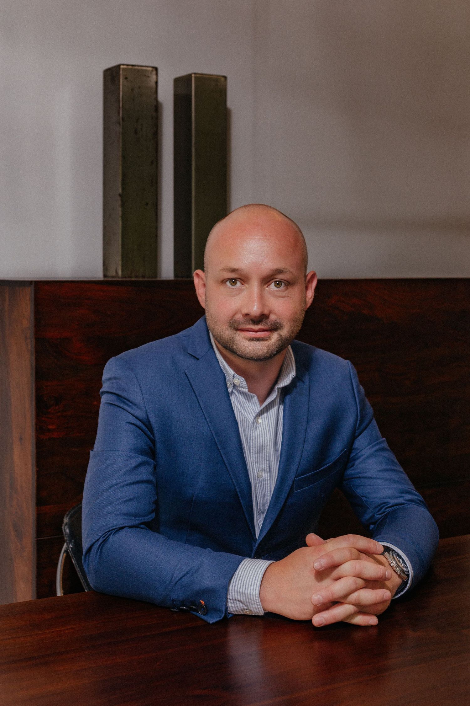 Bald man in blue blazer sits at wooden table with hands clasped in professional portrait setting.
