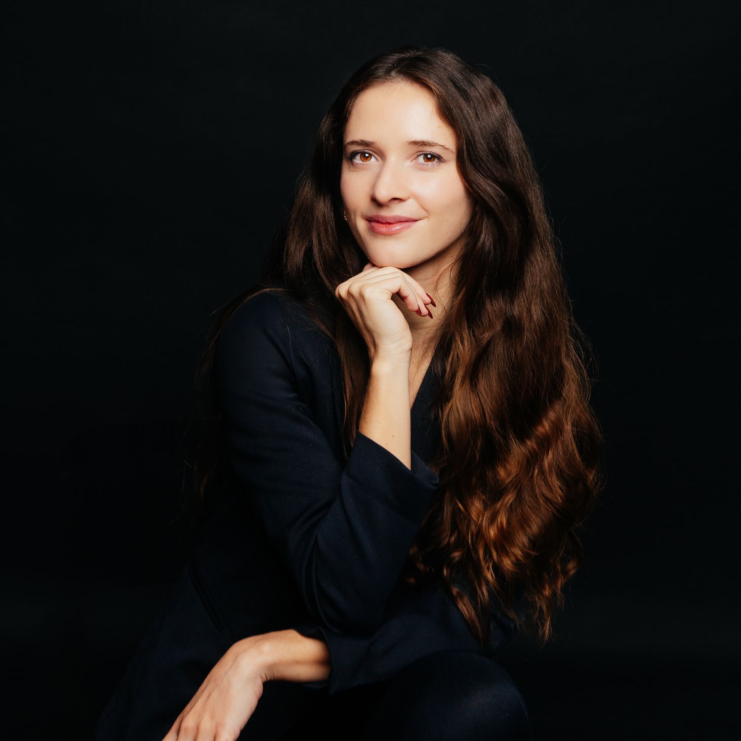 Young brunette woman in dark blazer poses thoughtfully with hand on chin against black background.