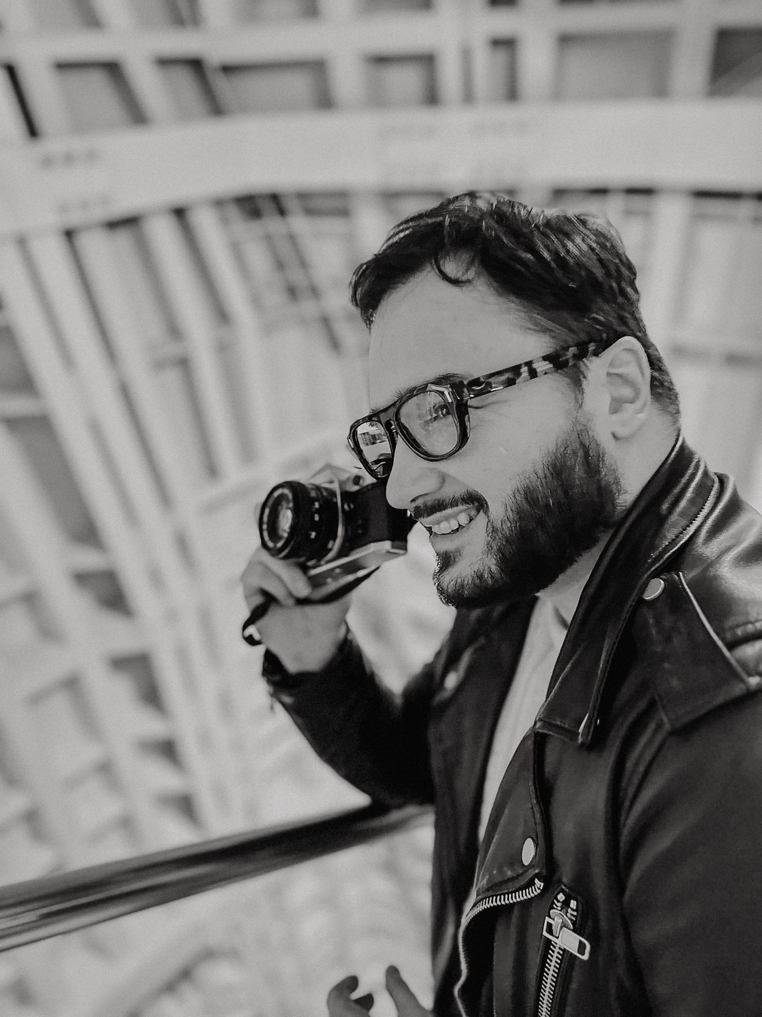Bearded man with glasses holding a vintage camera outdoors in black and white photography.