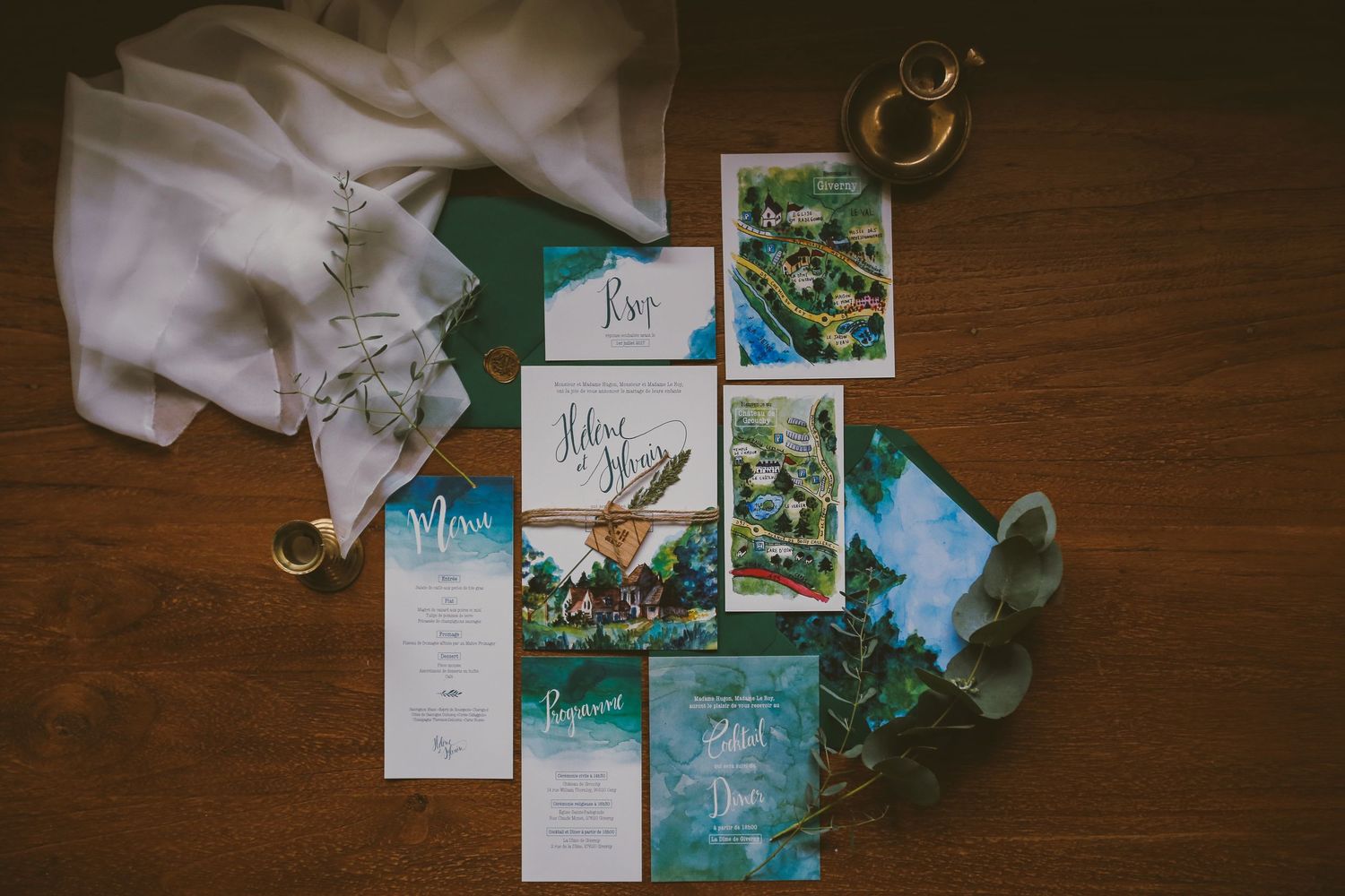 Elegant wedding invitation suite with watercolor designs in teal and green tones displayed on a wooden table.
