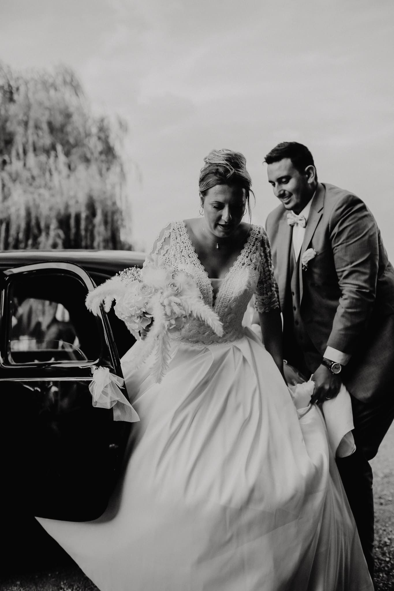 Black and white photo of groom helping bride exit a vintage car at an outdoor wedding.