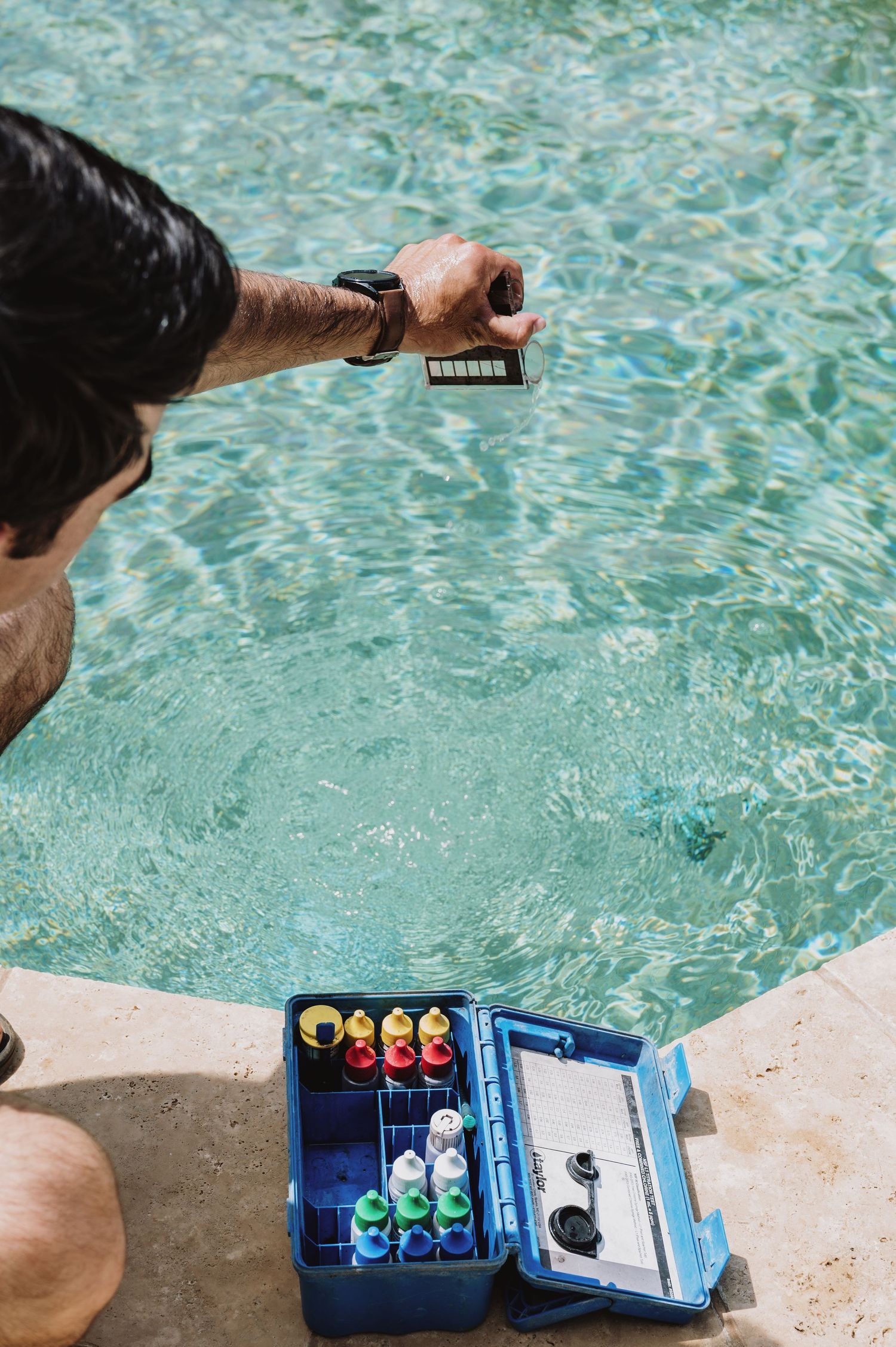Pool technician testing water chemistry with a test strip and colorful chemical kit beside a clear blue swimming pool.