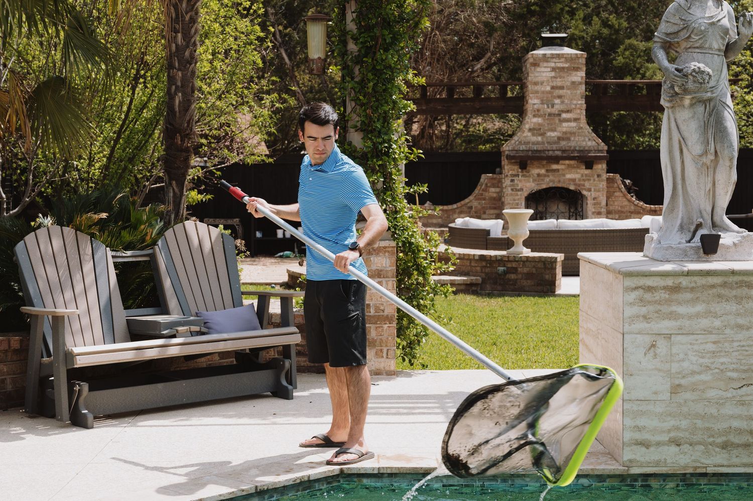 Man cleaning a backyard pool with a net skimmer, surrounded by patio chairs and stone fireplace.