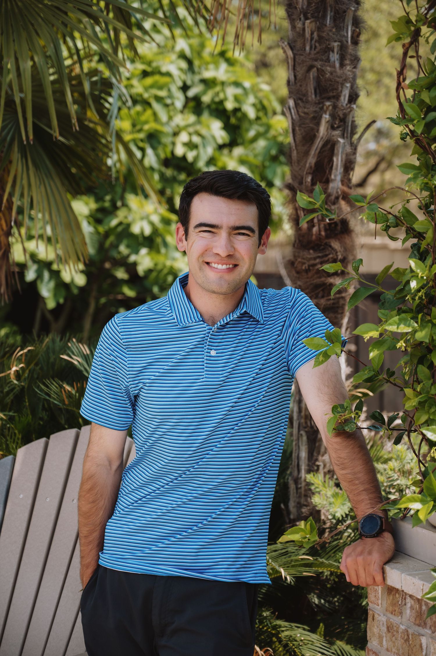 A smiling young pool tech in a blue striped polo shirt stands outdoors near tropical plants and a palm tree.