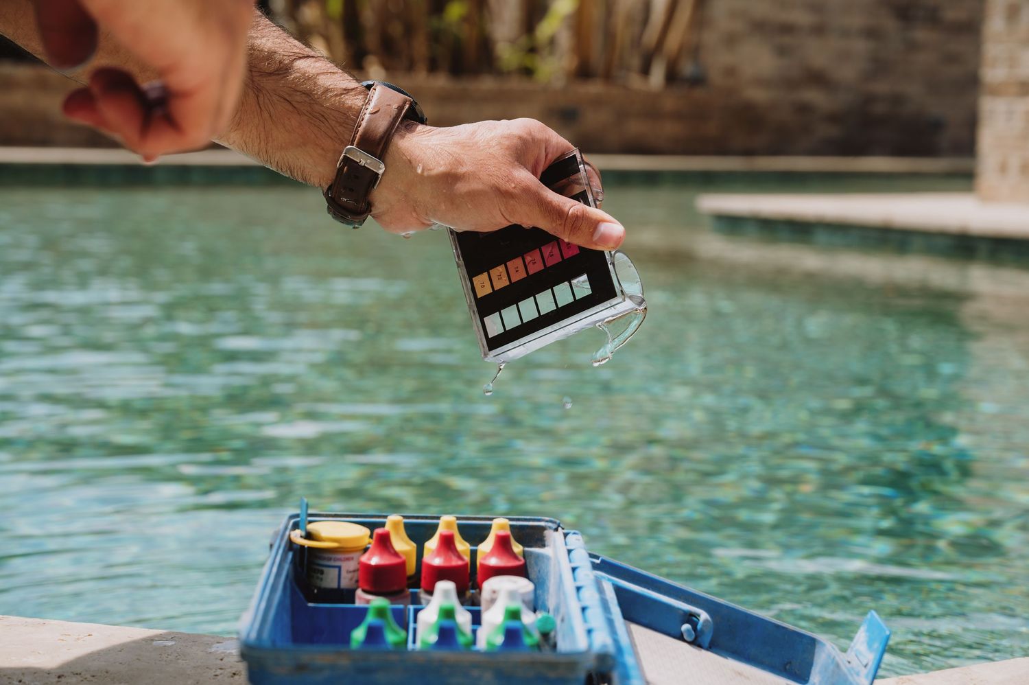 Person testing pool water chemistry using a colorimetric test kit with reagent bottles beside a clear blue swimming pool.