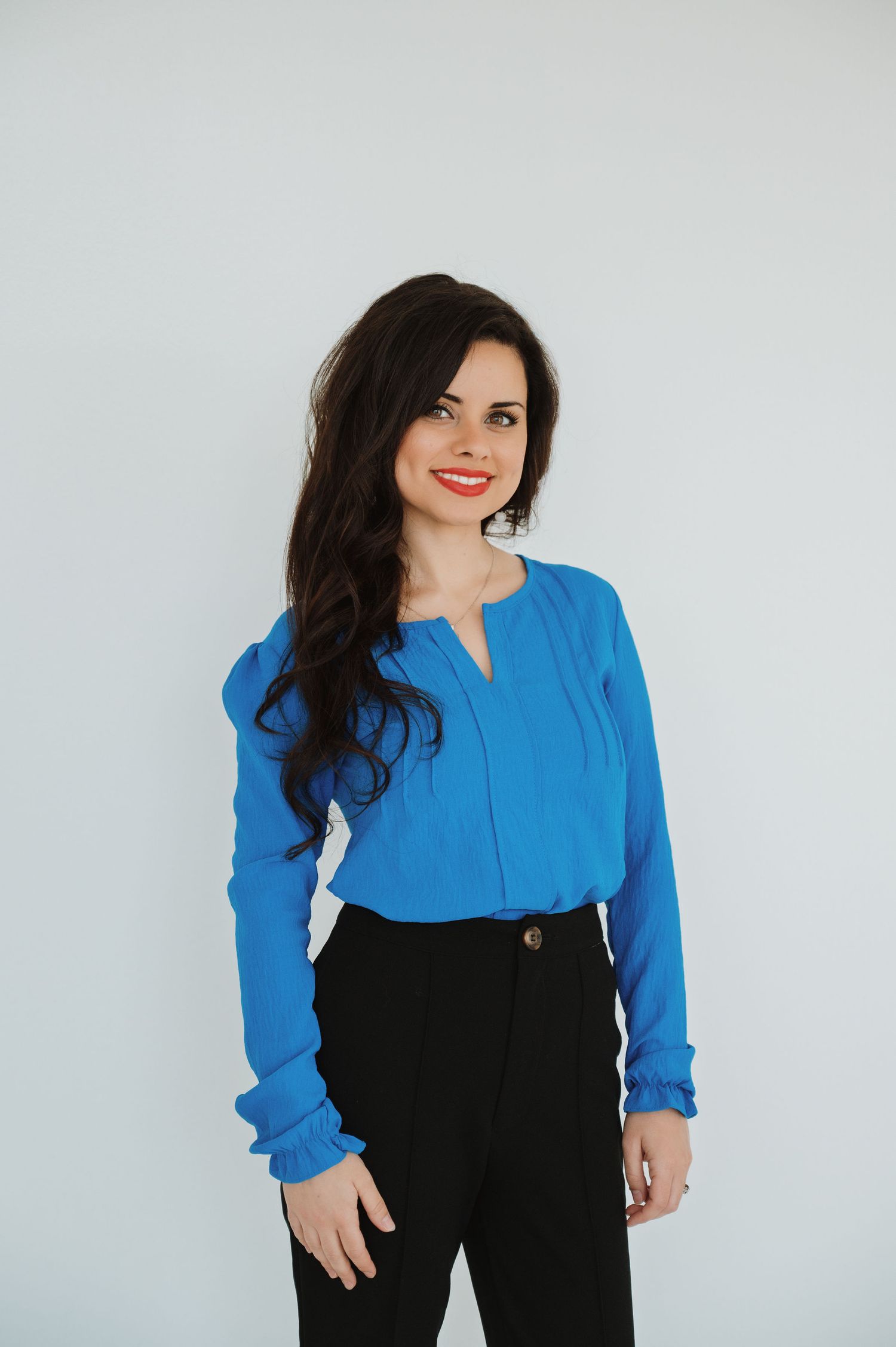 Woman in blue blouse and black pants smiling against white background.