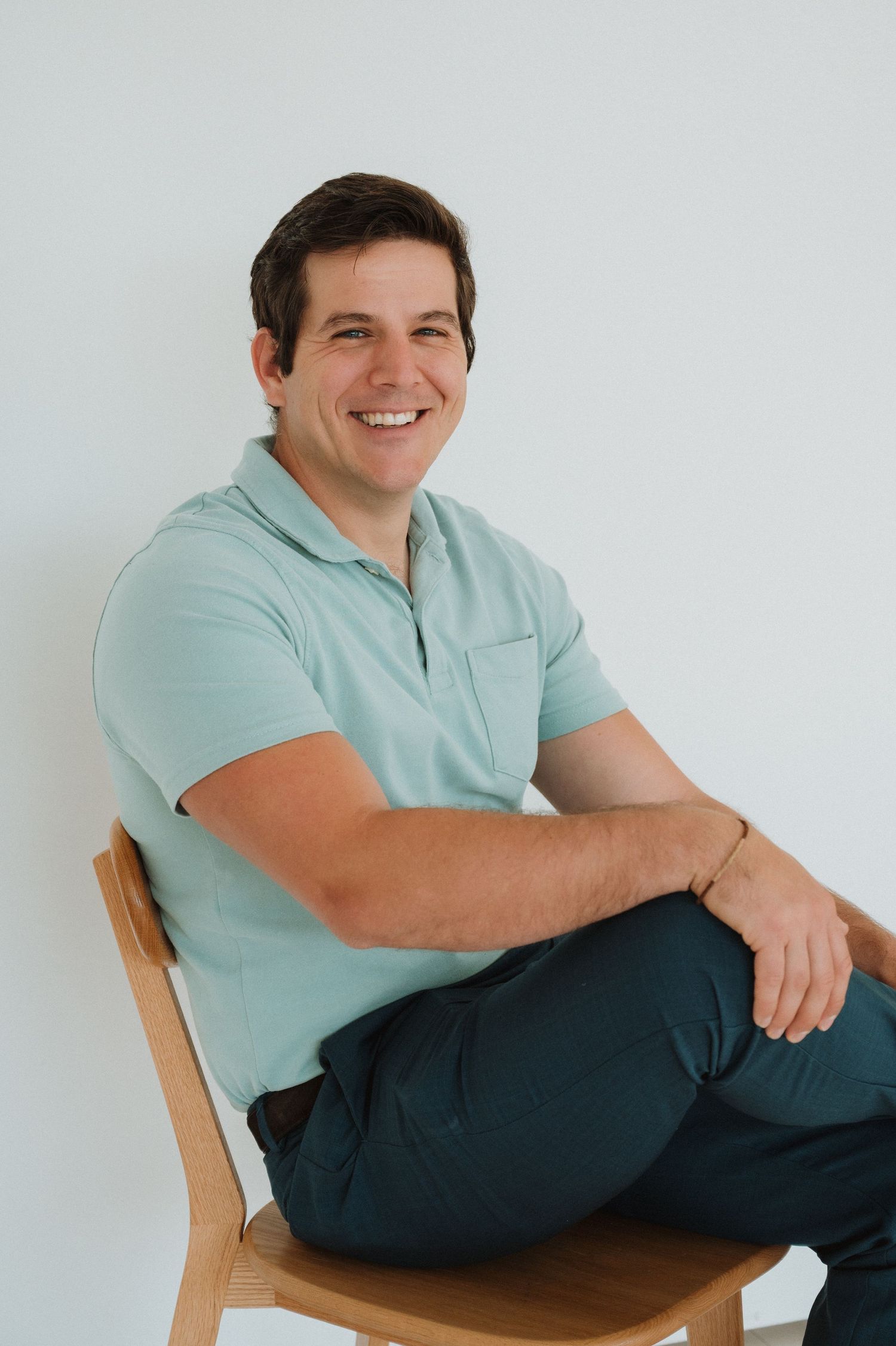 Smiling CPO Instructor in mint polo shirt sitting relaxed on wooden chair against white background.