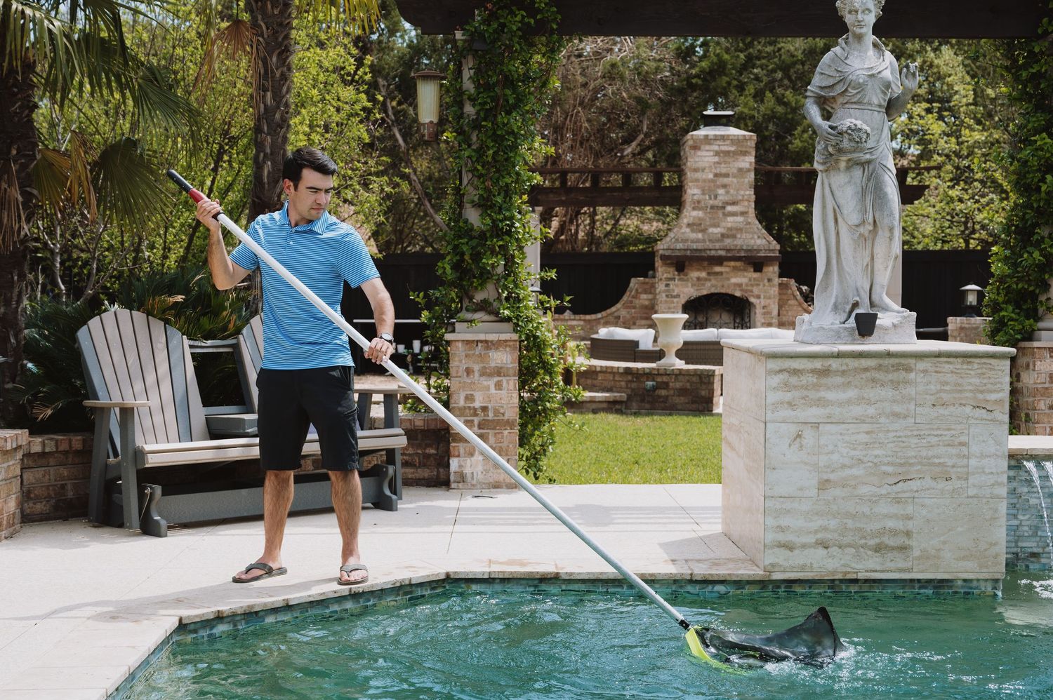 A pool technician in a blue shirt uses a net skimmer to clean a backyard swimming pool near a stone statue.