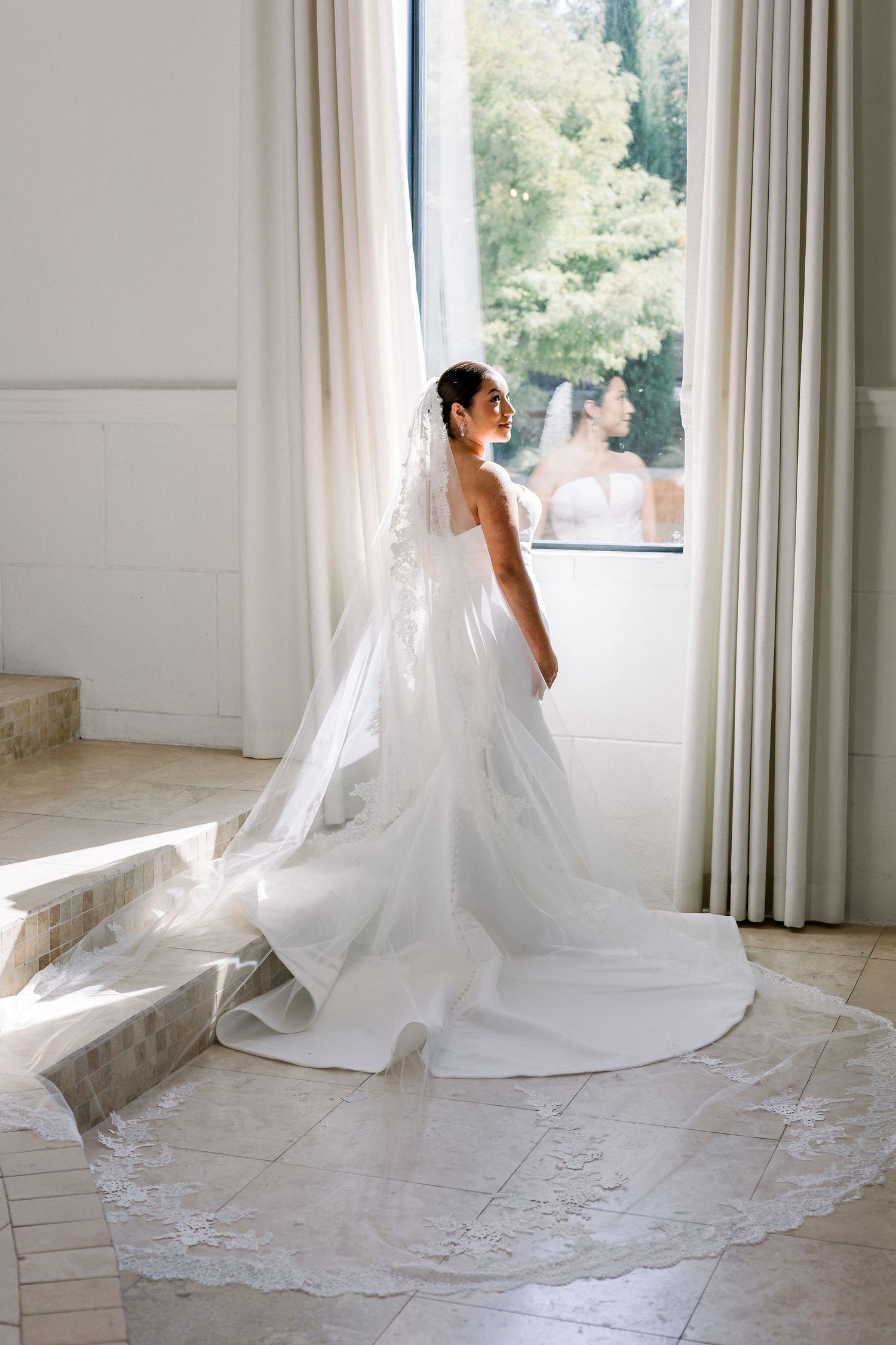 Bride in a stunning white lace-trimmed cathedral veil and strapless gown standing by a sunlit window.