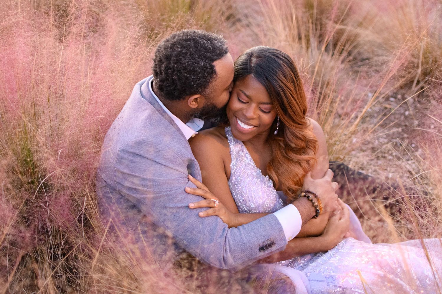 A couple shares a romantic embrace among pink pampas grass, the man kissing the woman's cheek as she smiles joyfully.