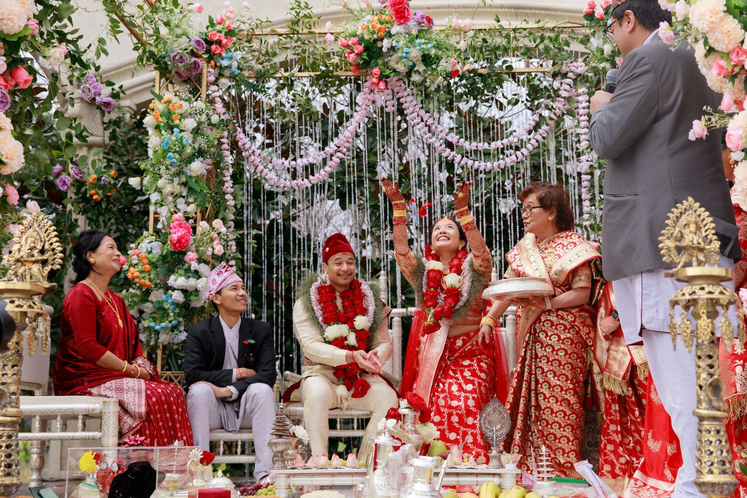 Colorful Hindu wedding ceremony with bride and groom in red attire seated under floral mandap with family members.
