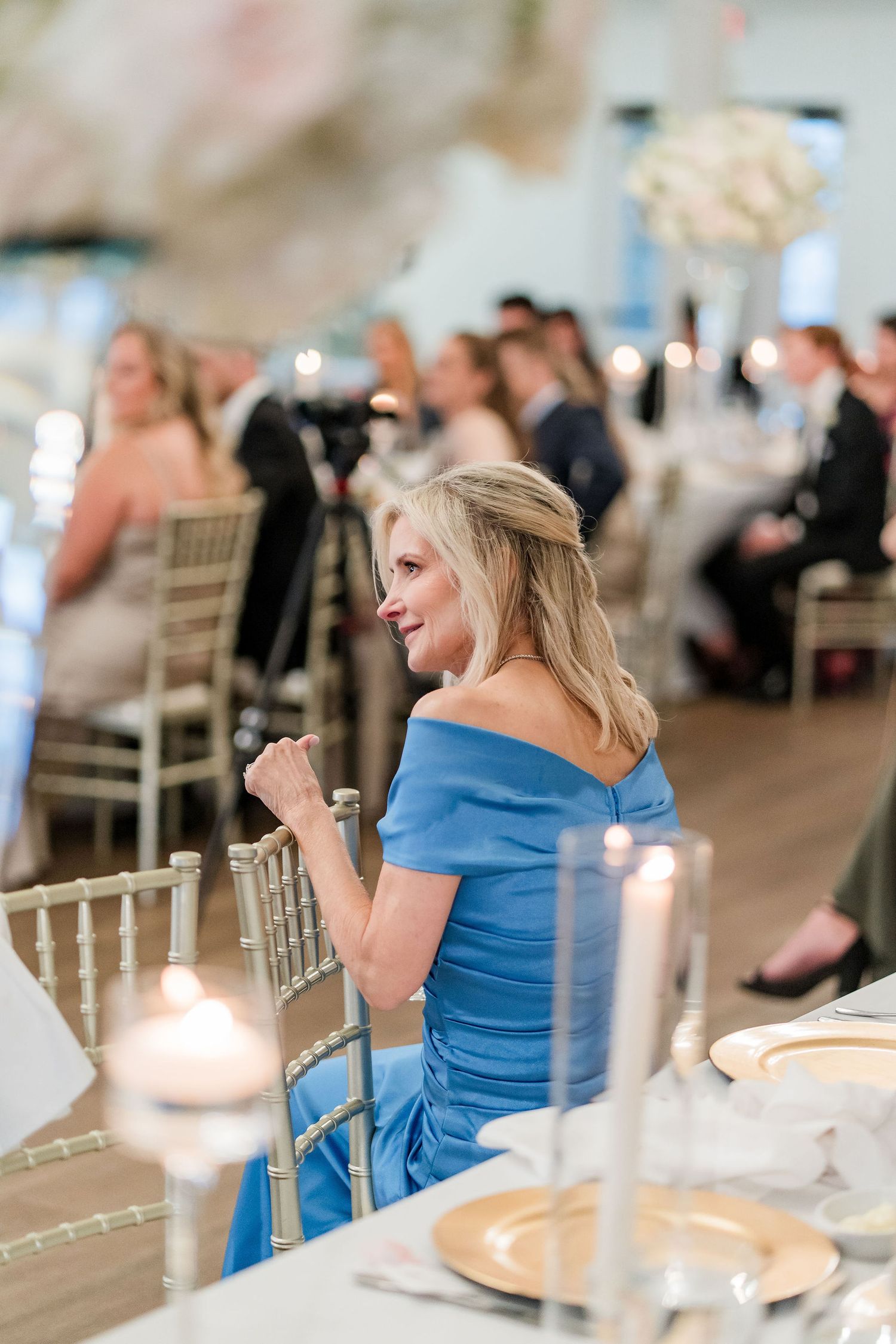 Woman in blue off-shoulder gown seated at elegant wedding reception table with gold chargers and candles.