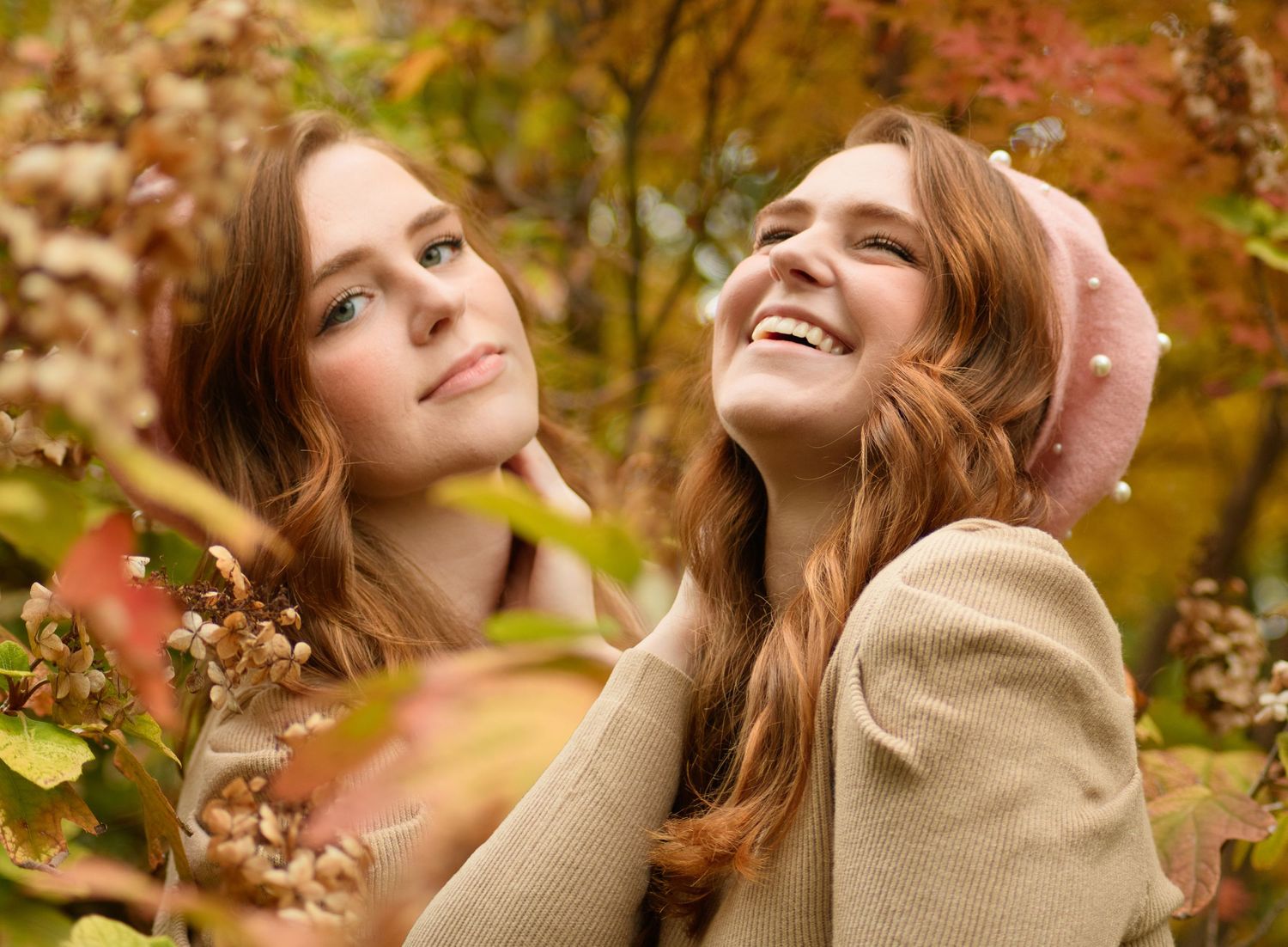 Two smiling young women with auburn hair enjoy autumn foliage surrounded by colorful fall leaves outdoors.