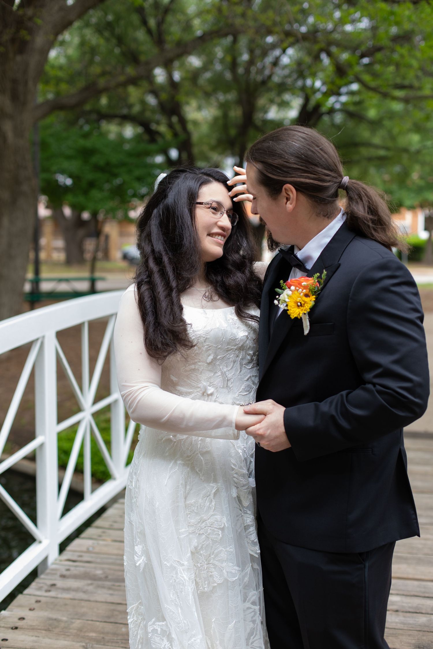 A couple shares a romantic moment on a white bridge in a garden setting wearing wedding attire.