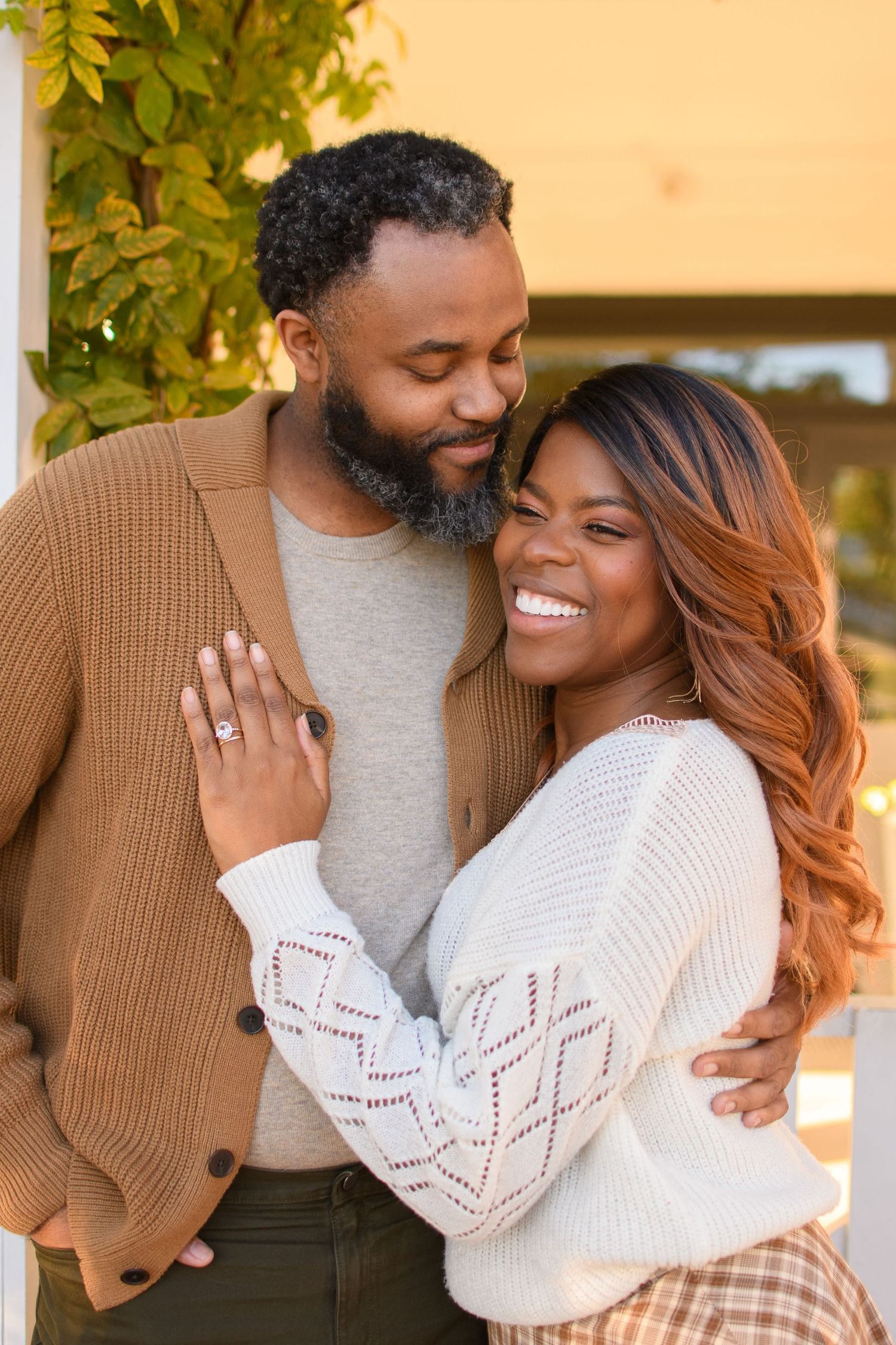 A smiling woman with an engagement ring embraces her bearded partner in a warm outdoor autumn setting.