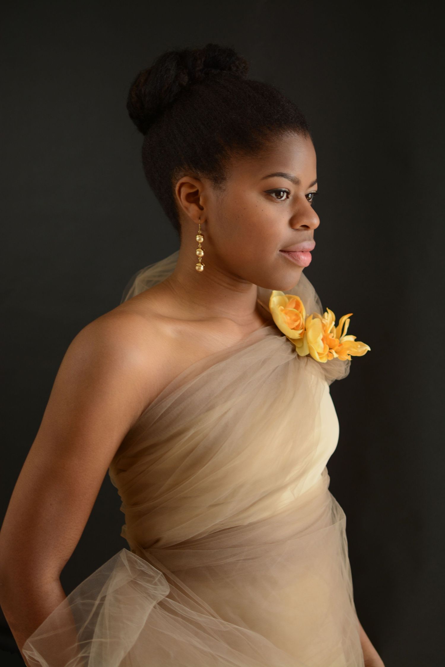 Elegant Black woman in beige tulle one-shoulder gown with yellow flowers poses on dark background.
