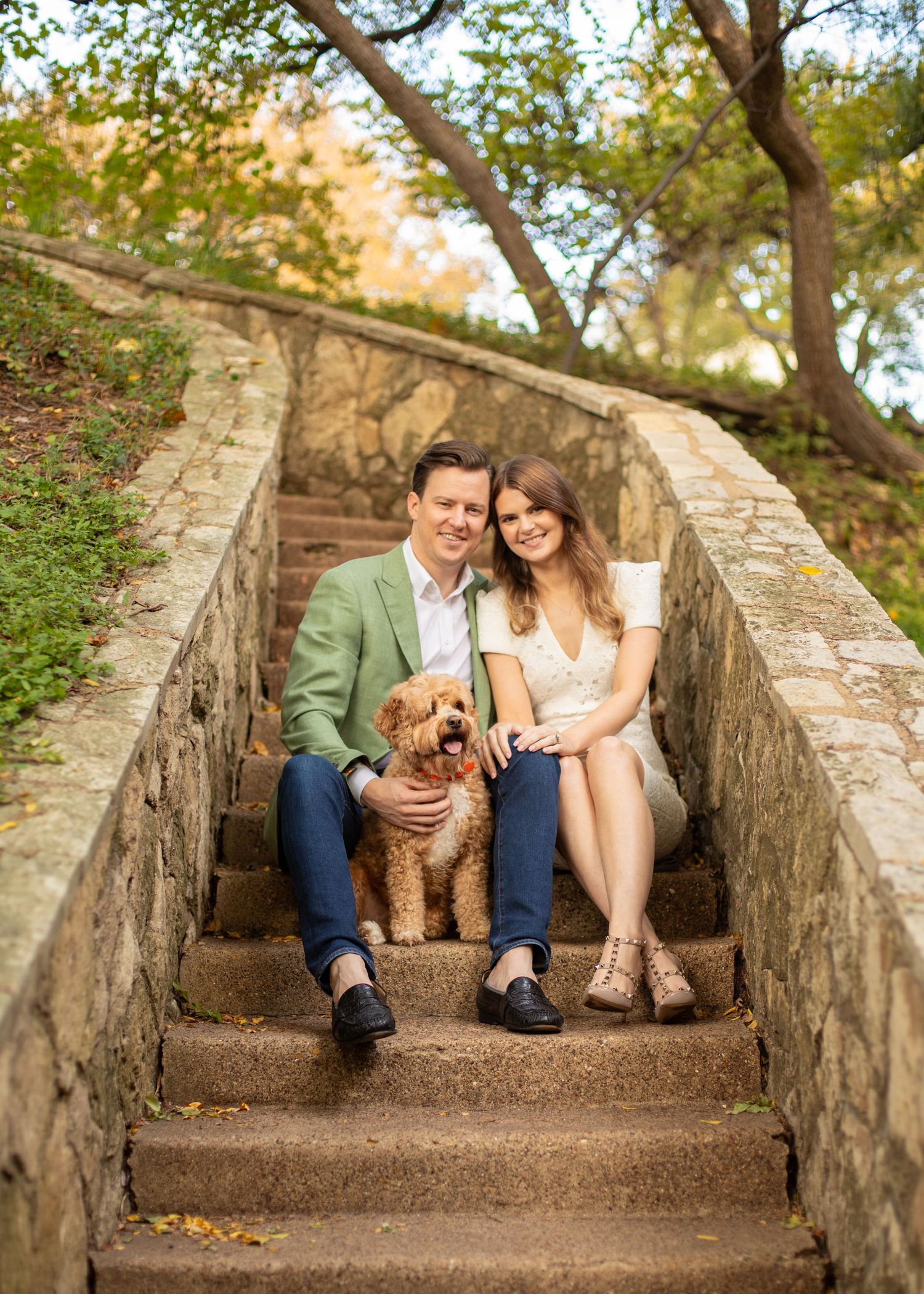 Happy couple posing with golden doodle dog on stone staircase in autumn park.