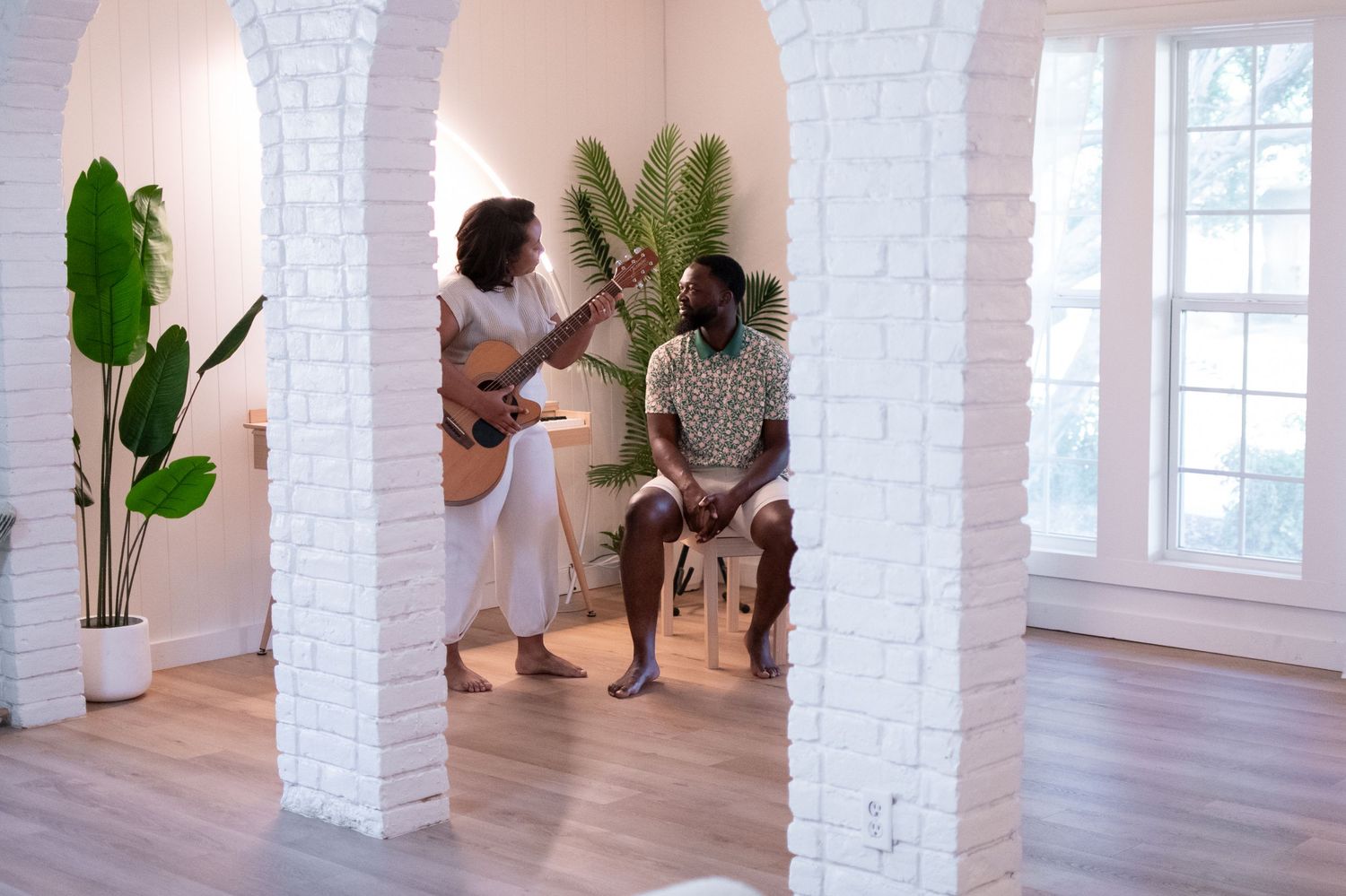 Couple sharing musical moment with guitar in sunlit white brick archway room.