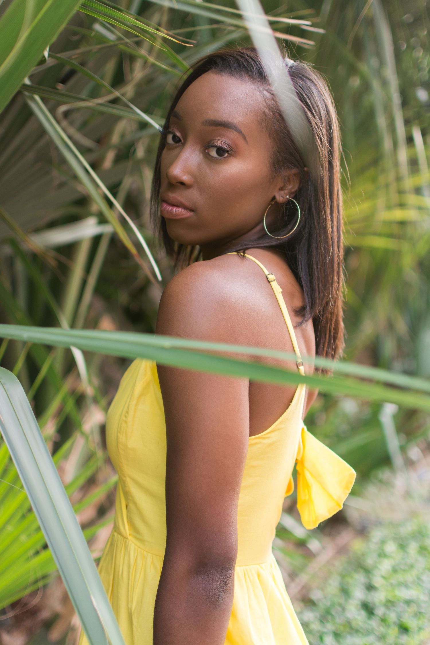 Young Black woman in yellow sundress poses among tropical palm fronds outdoors.