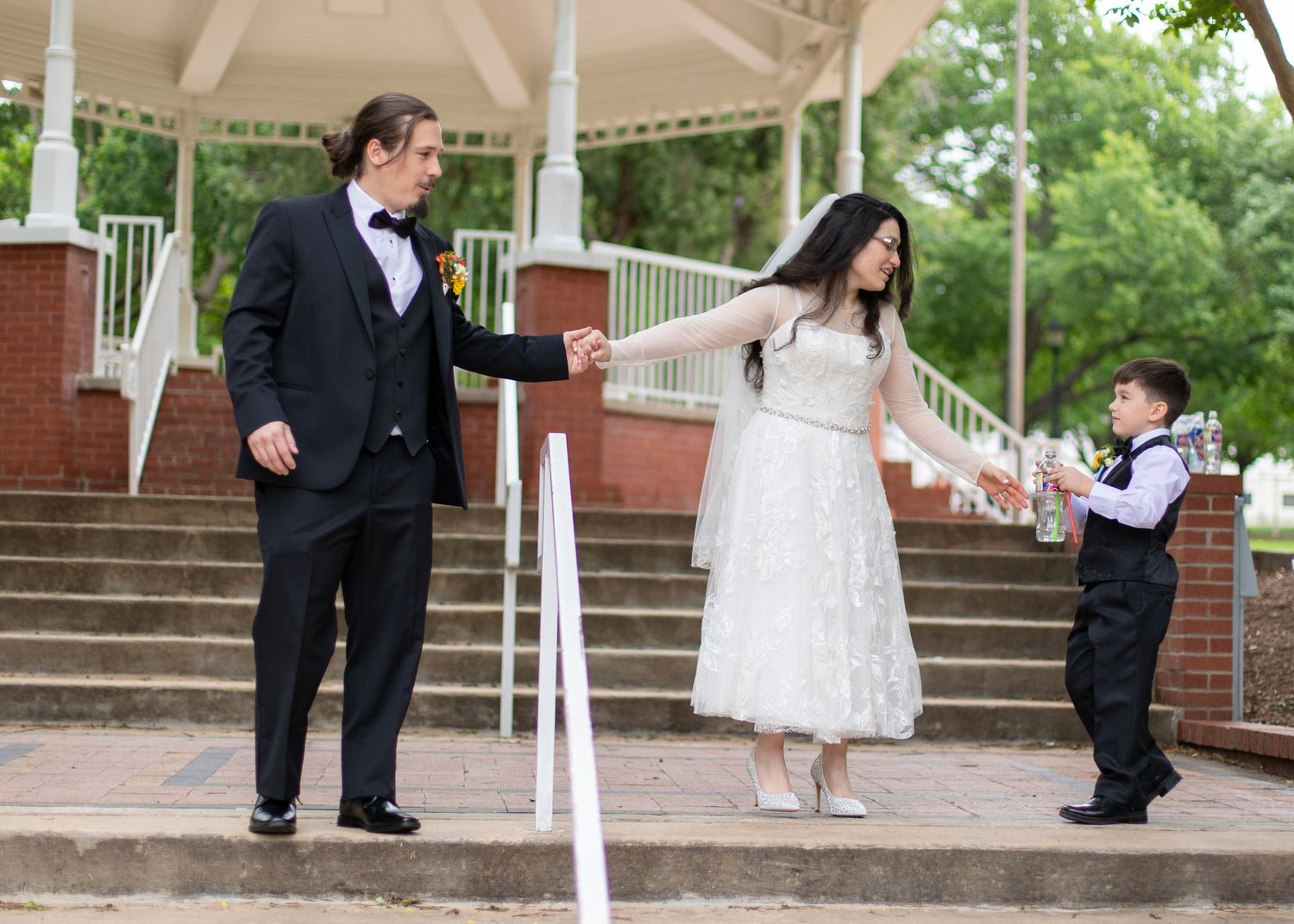 Wedding party members pose playfully on brick steps under white gazebo.