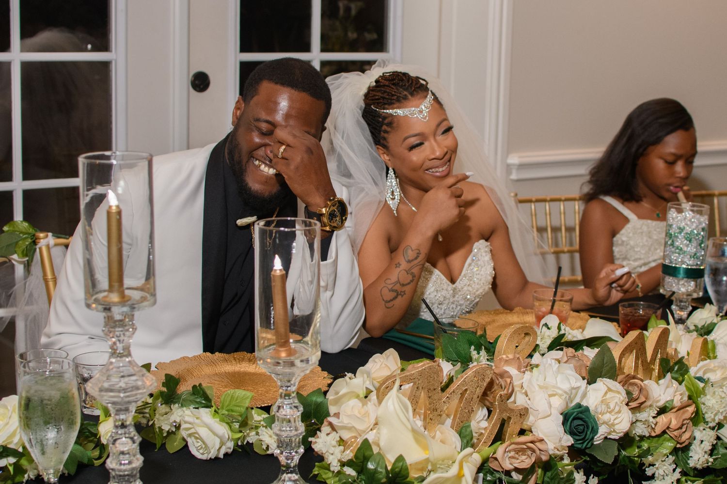 Smiling bride and groom share a tender moment together at floral-decorated wedding sweetheart table.