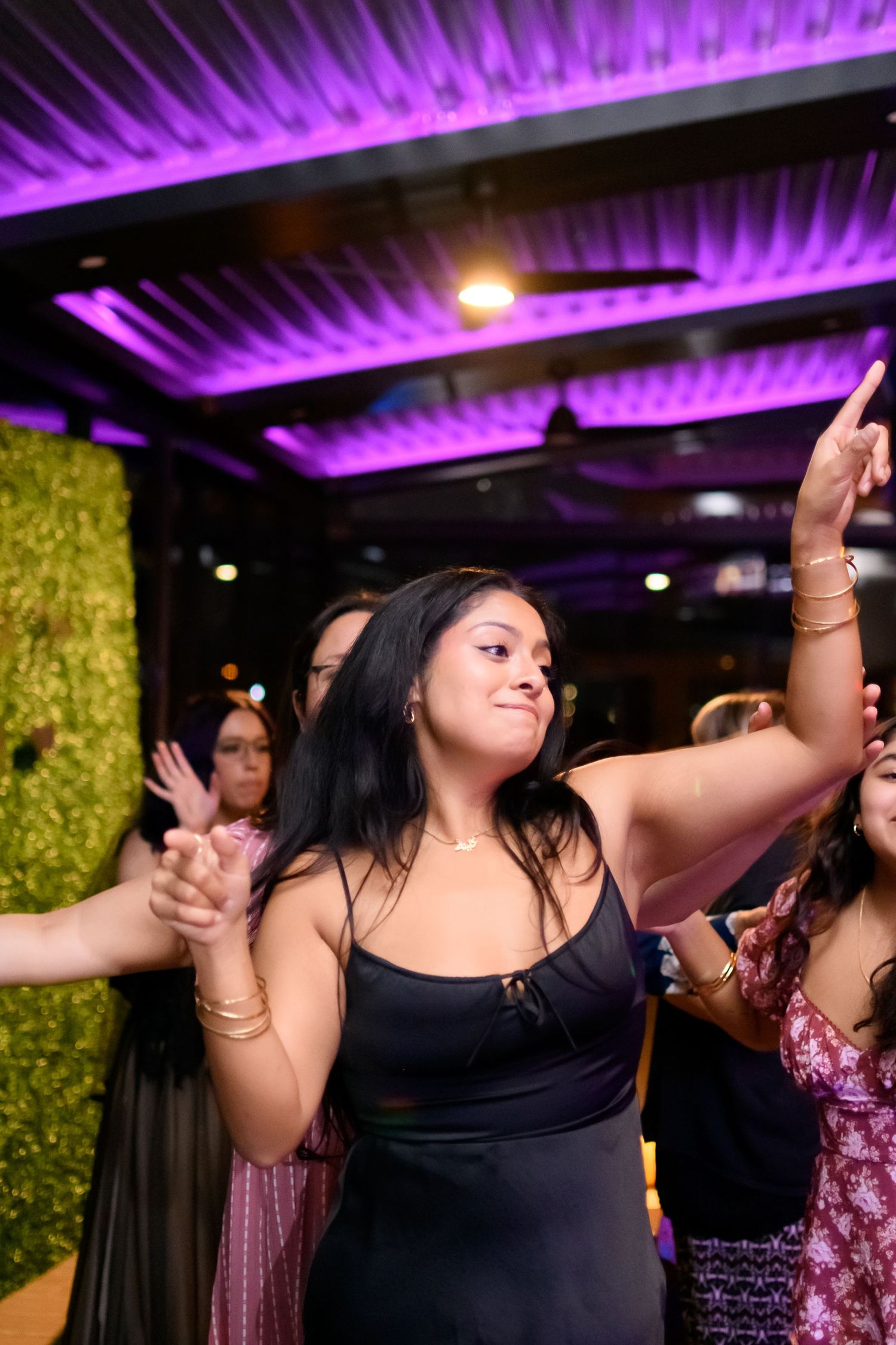 Young woman in black dress dancing joyfully with arm raised under purple lights at wedding reception.