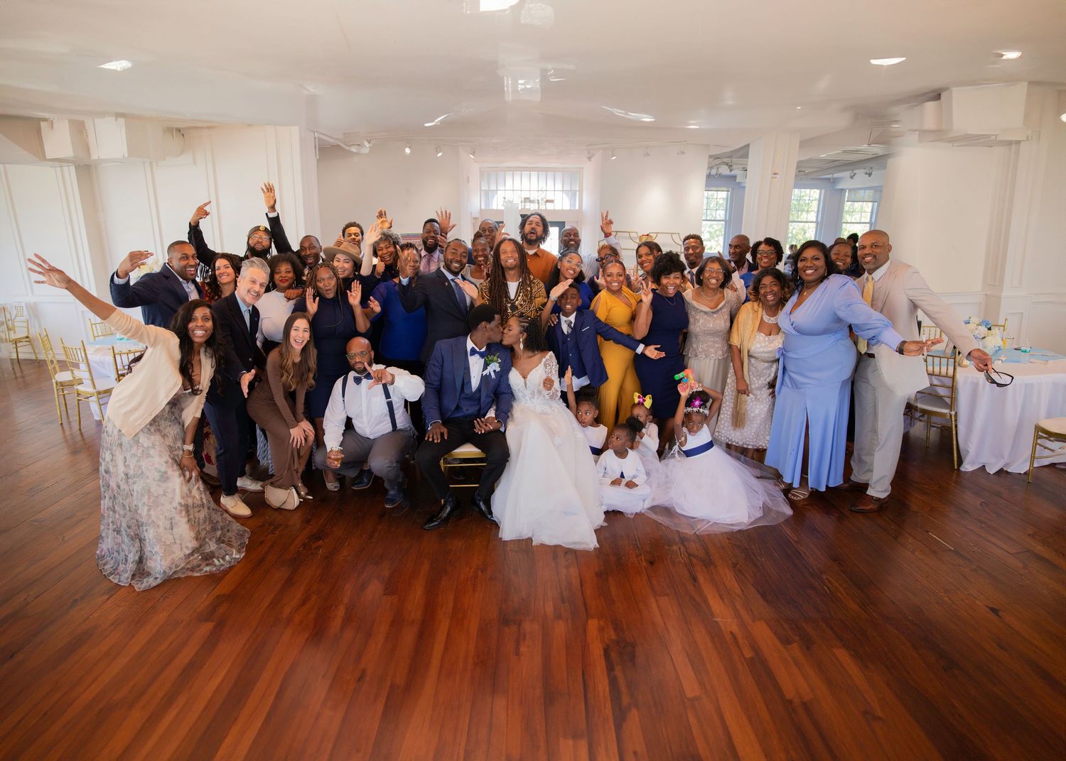 Large wedding group photo with bride and groom surrounded by joyful guests celebrating in elegant white reception hall.