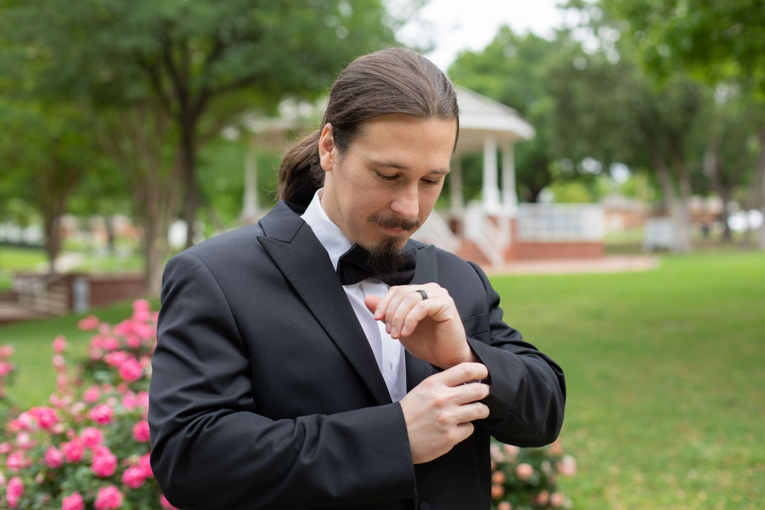 A person in a black suit adjusts their tie outdoors with pink flowers and a gazebo in the background.