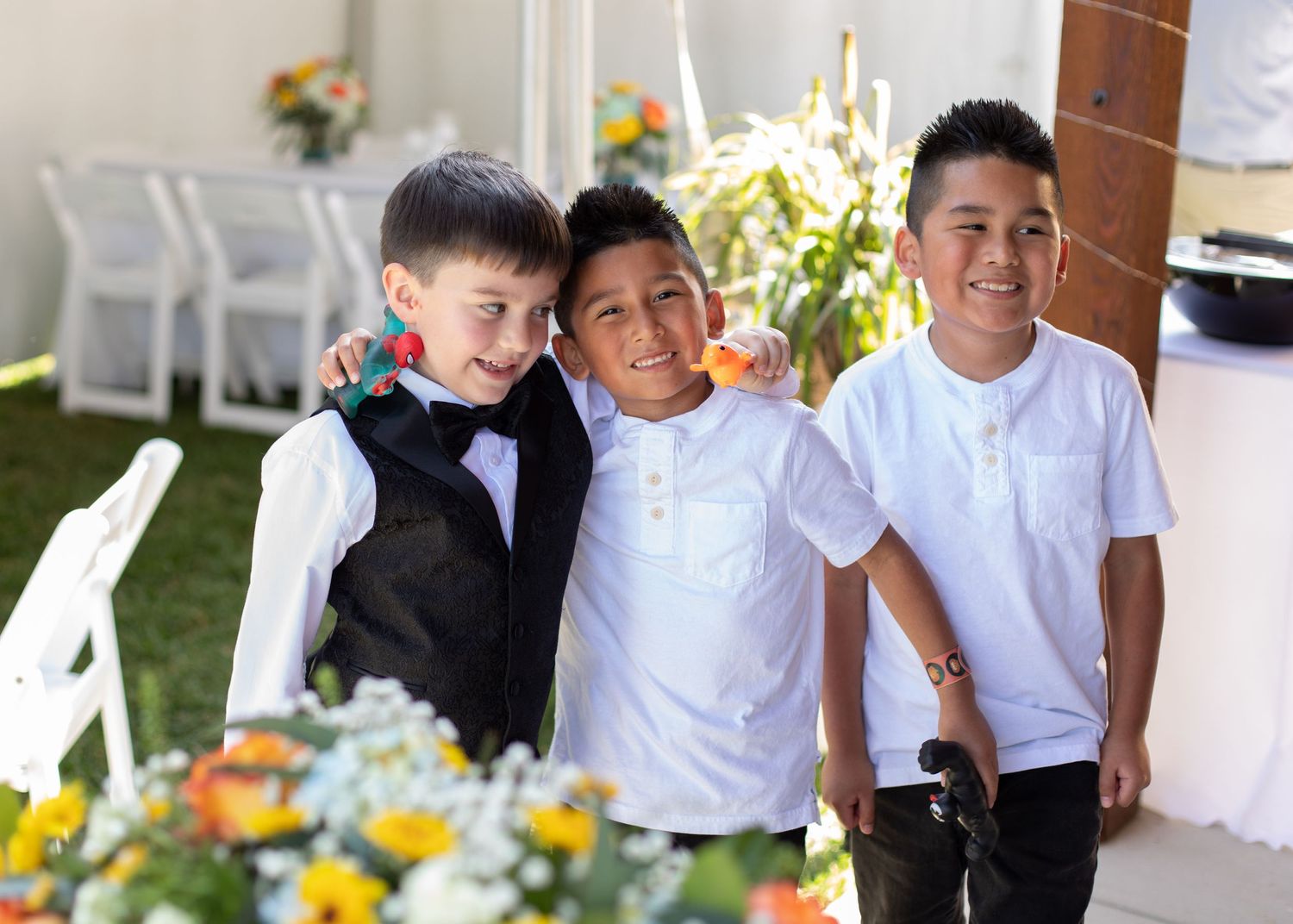 Three young boys in formal attire smiling together at an outdoor event.