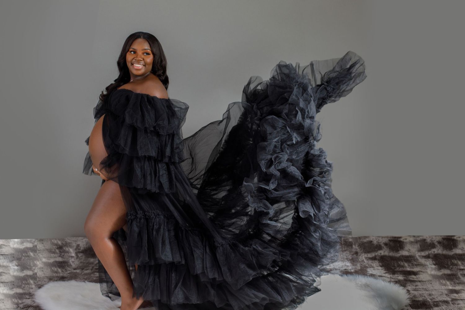 Pregnant Black woman in black ruffled tulle gown smiles joyfully while dress flows around her.