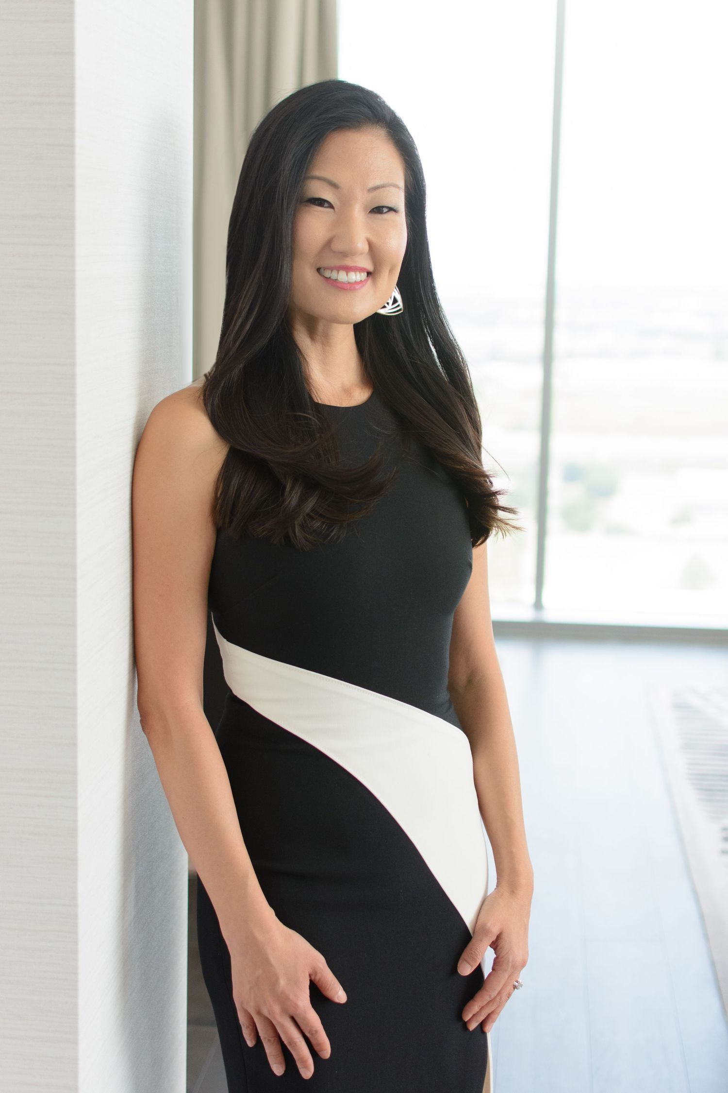 Elegant Asian woman in a black and white fitted dress smiles while posing near a bright window indoors.
