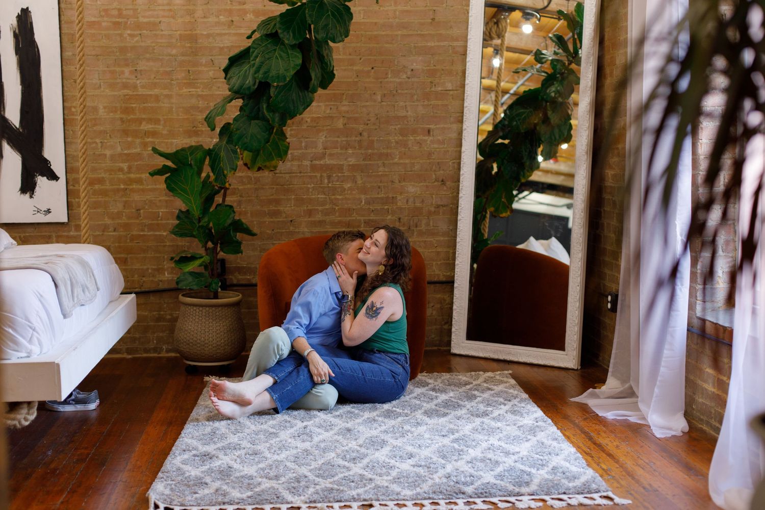 Couple sharing a kiss while sitting on a rug in a cozy room with brick walls and lush green plants.