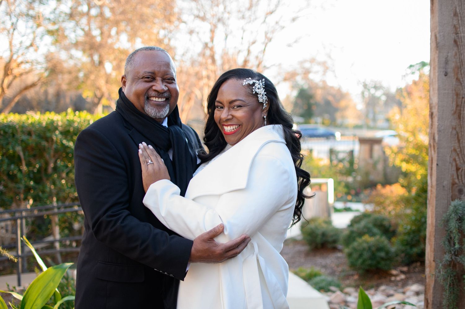 Smiling couple embracing outdoors, the woman in a white coat with a floral hair accessory and the man in a dark coat, among autumn trees.