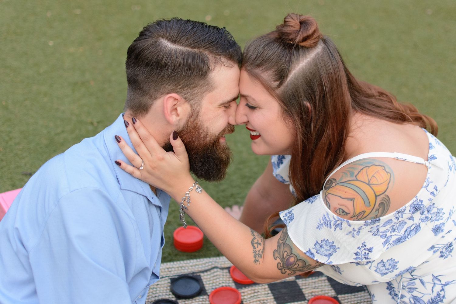 Bearded man and tattooed woman share a romantic forehead touch while playing giant checkers on a lawn.