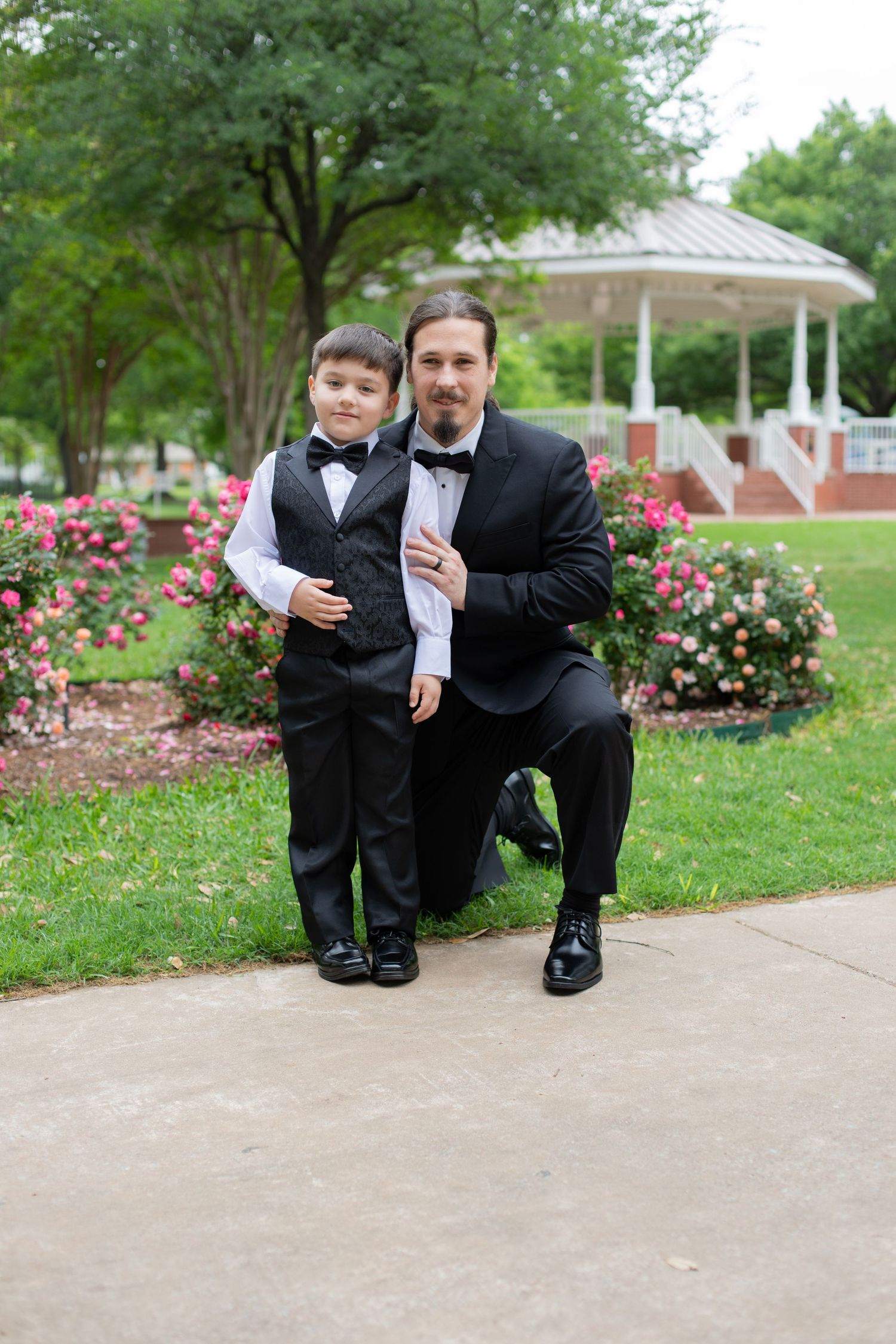 Two people in formal attire pose together on a path near blooming rose bushes and a gazebo.
