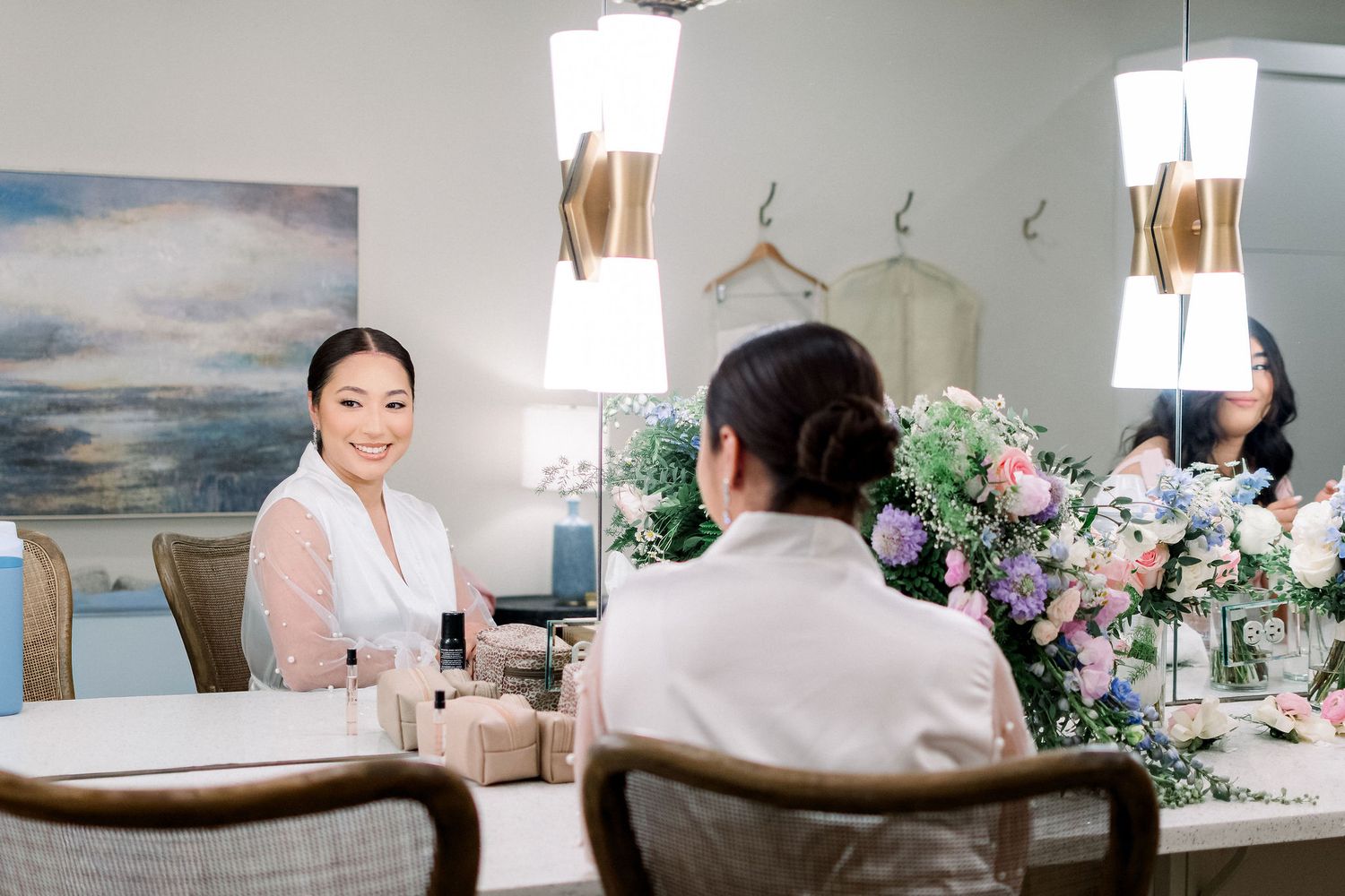 A smiling bride sits at a vanity mirror surrounded by pink and purple floral arrangements during wedding preparations.