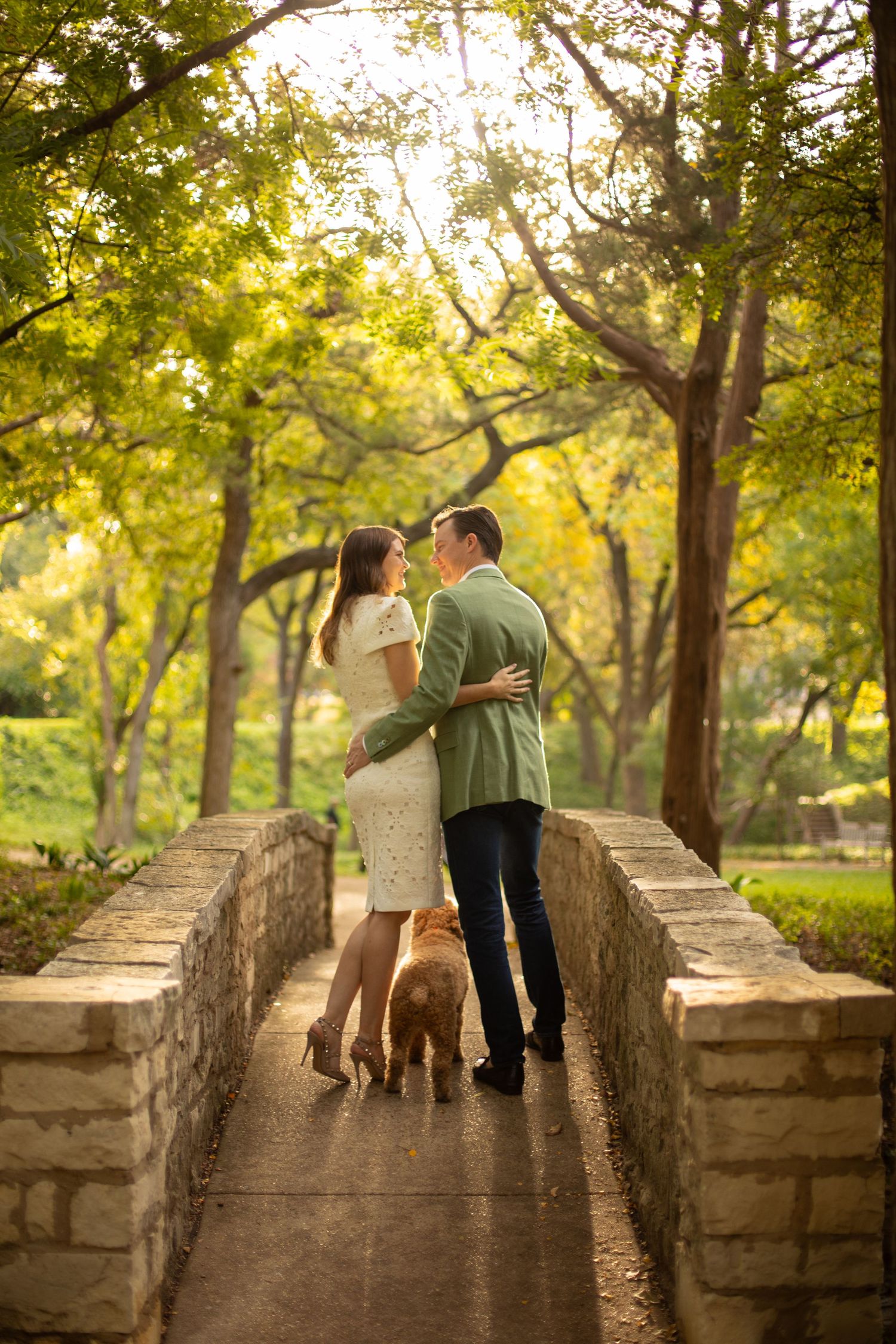 Couple embracing on stone bridge with golden dog amid golden autumn park trees.