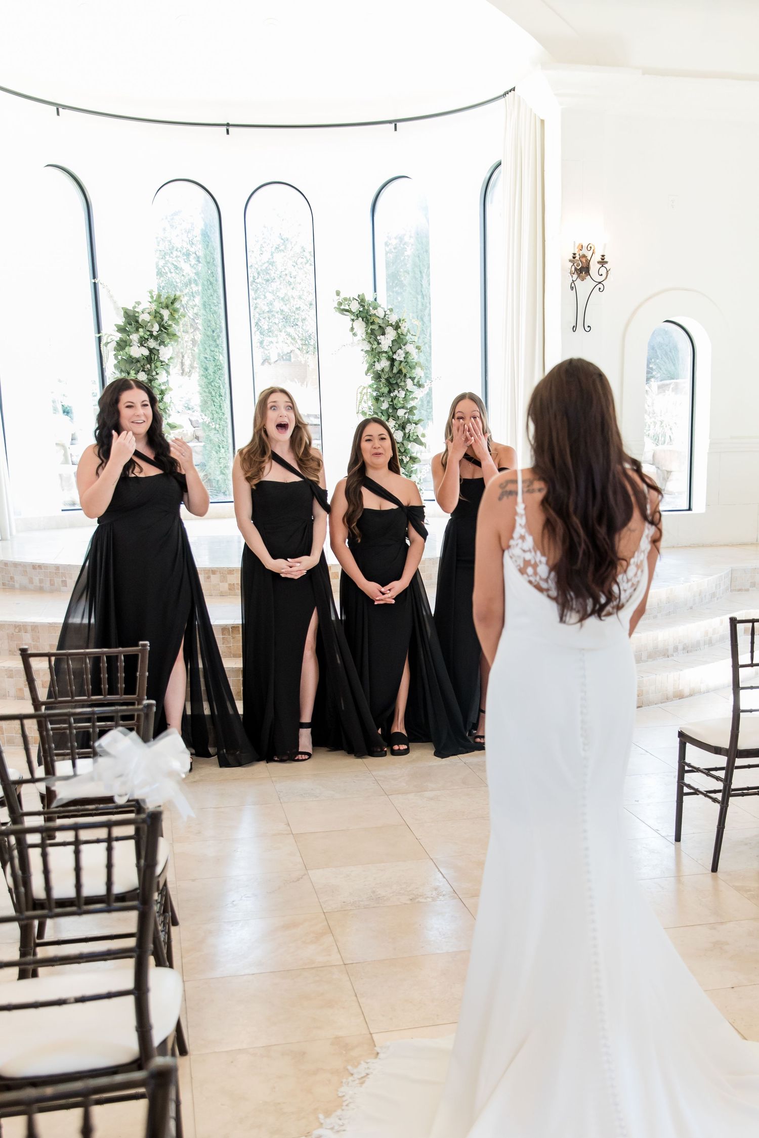 Bridesmaids in black gowns react with joy seeing the bride in her wedding dress for the first time.