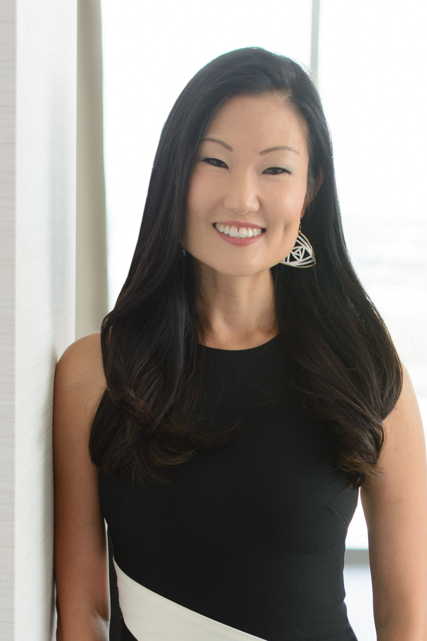 Smiling Asian woman with long dark hair wearing a black and white sleeveless dress leans against a wall.