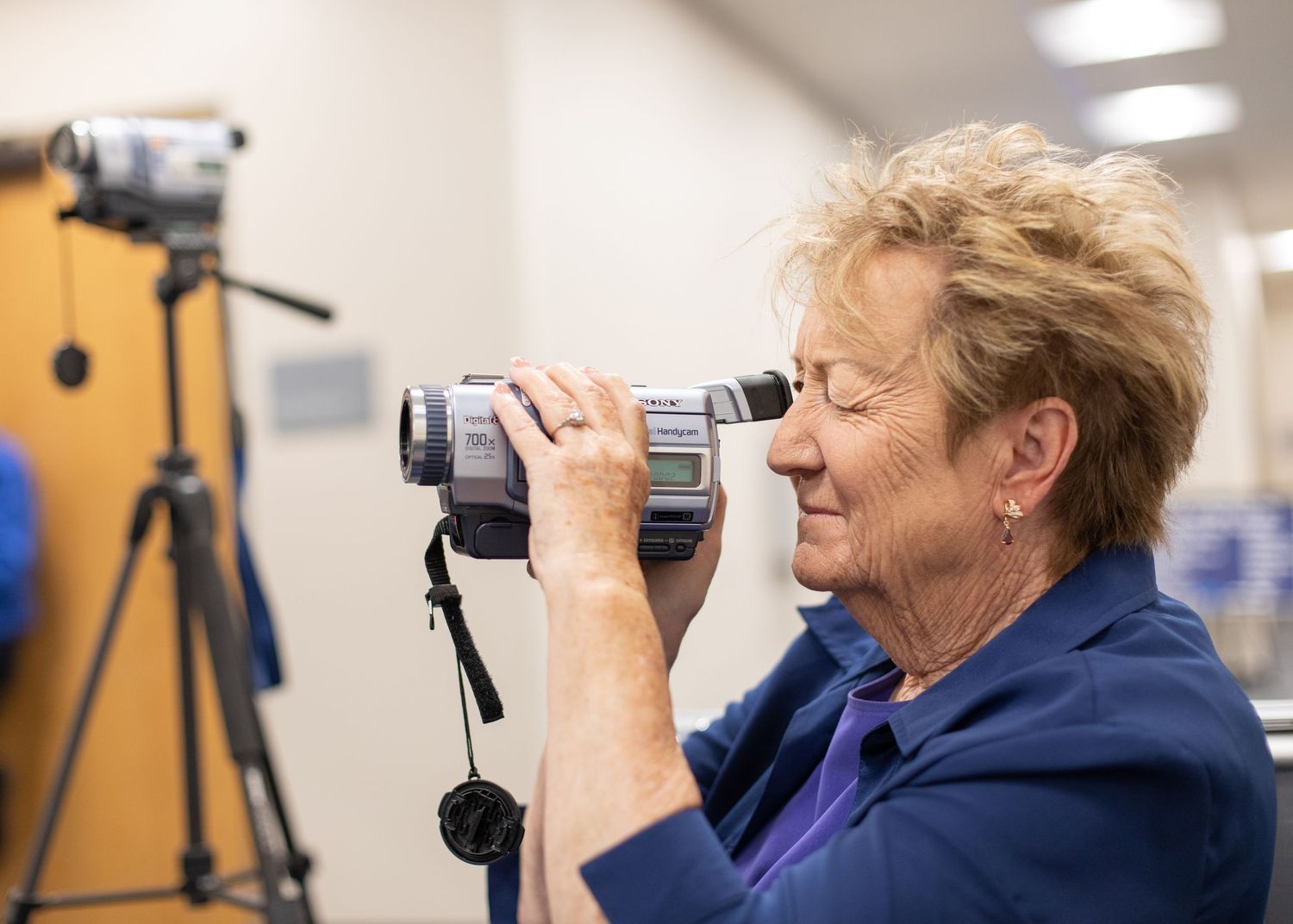 A person in blue using a camera while smiling in an indoor setting with photography equipment.