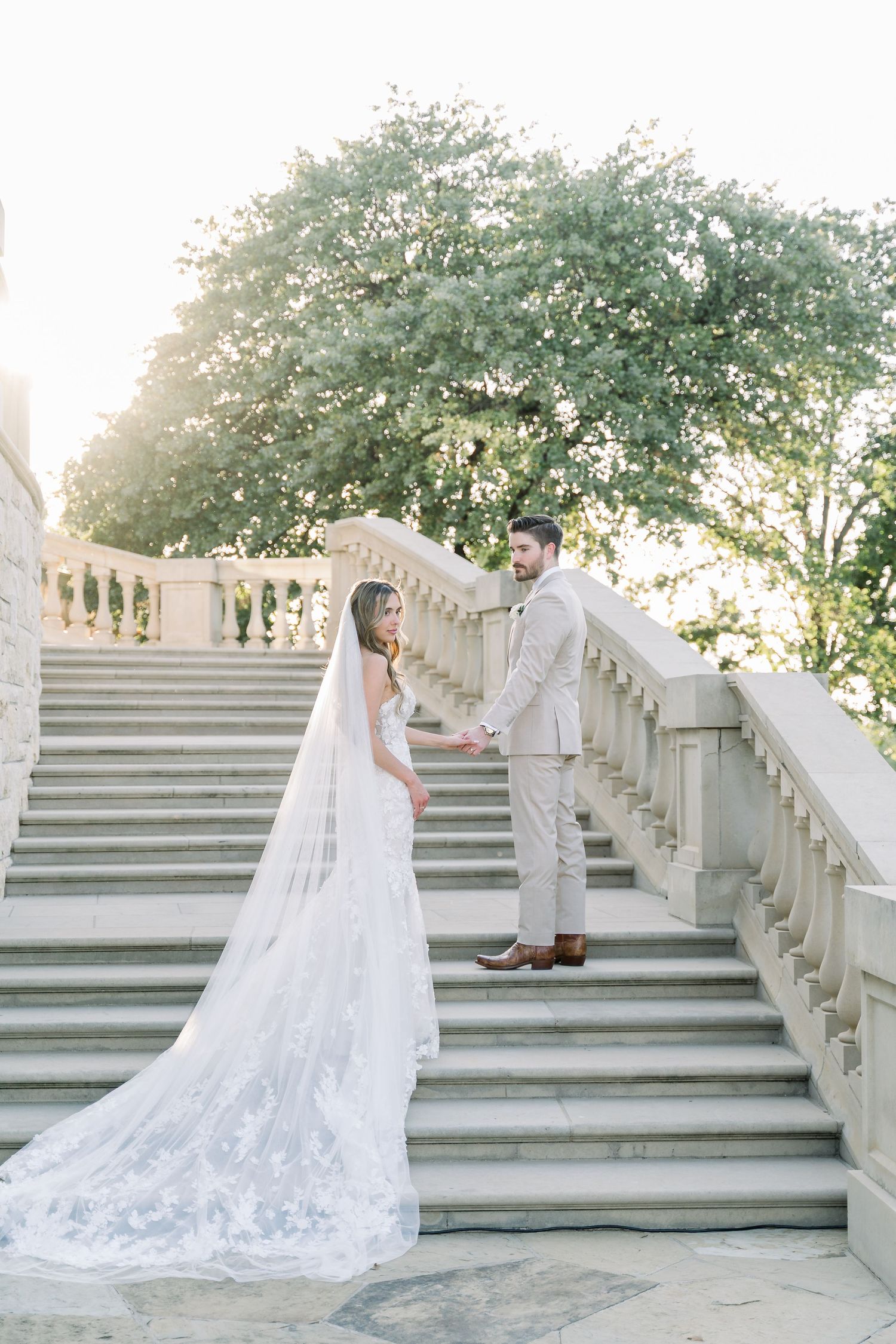 Bride and groom holding hands on grand stone staircase with flowing cathedral veil at golden hour.
