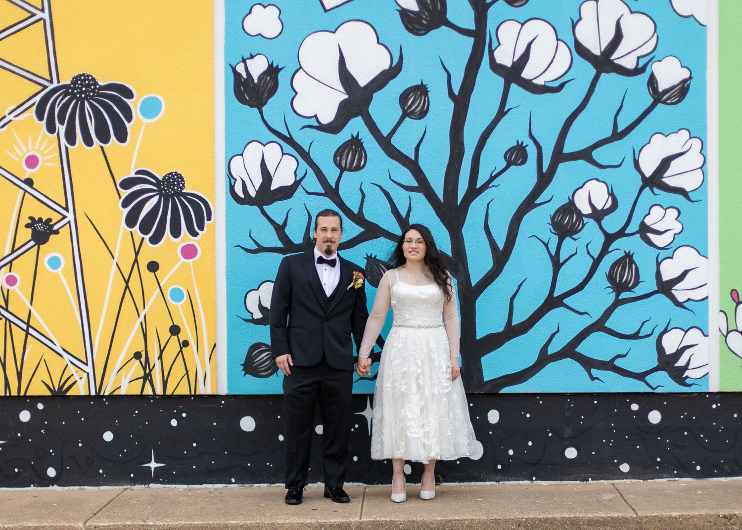 A couple poses in formal attire against a bright mural with cotton plants and yellow geometric designs.