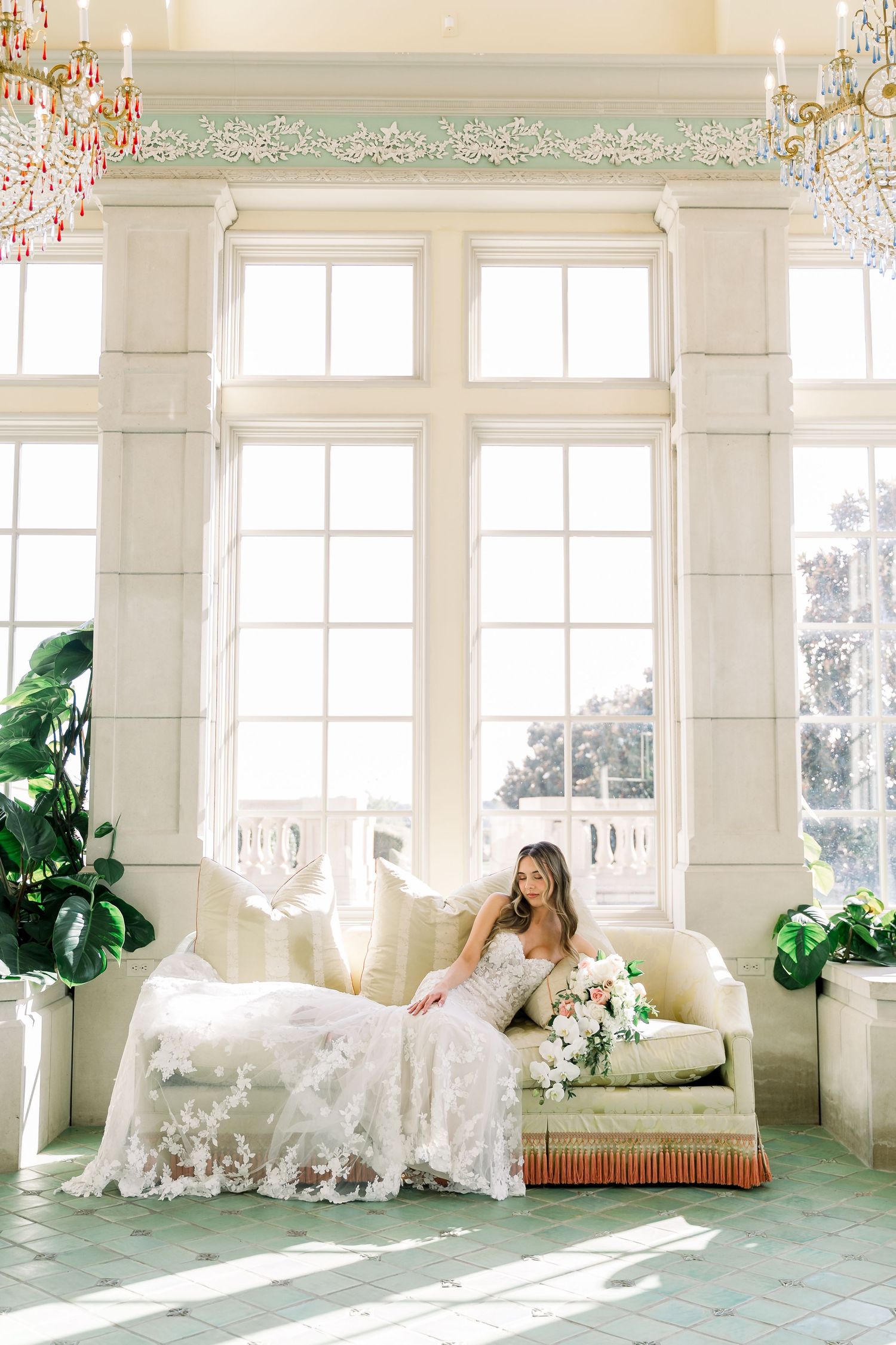 Bride in lace gown seated on vintage sofa in bright sunlit room with tall windows and chandeliers.